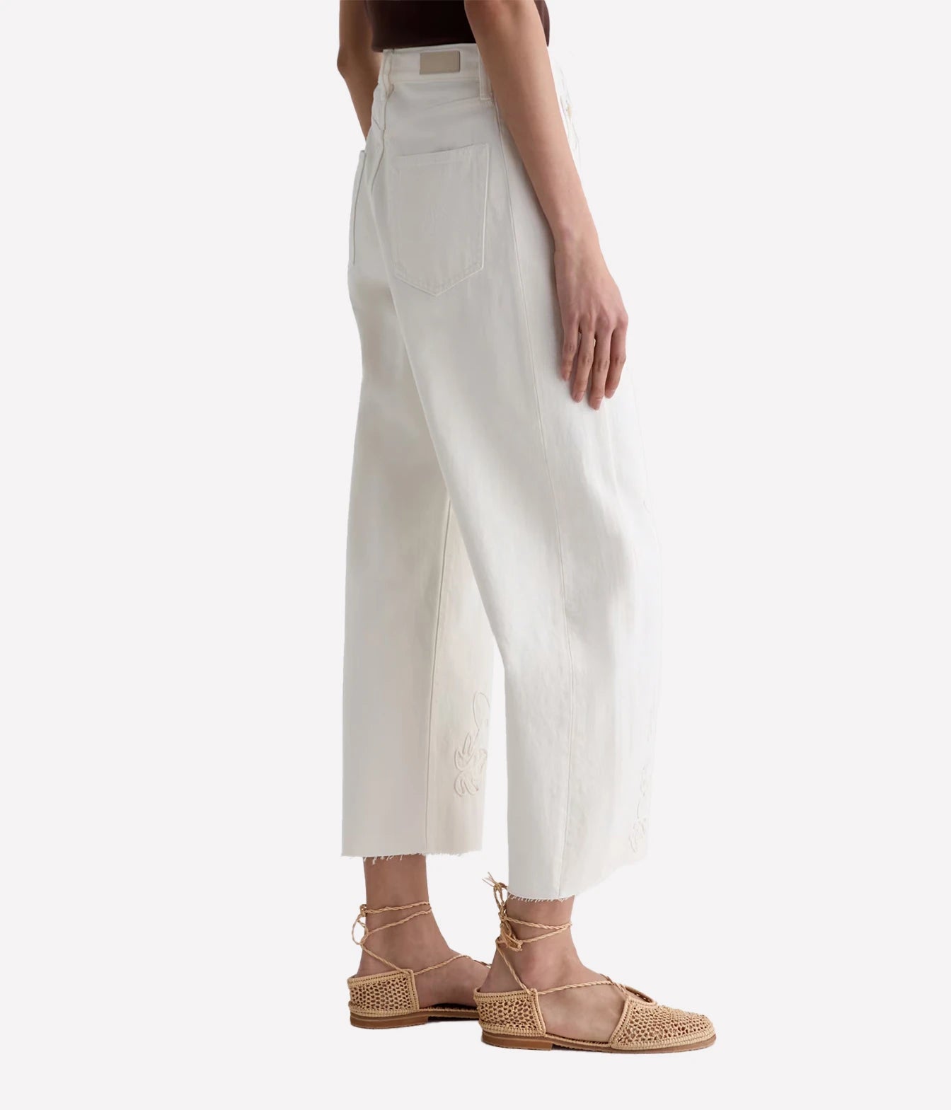 A model is shown from the side wearing high-waisted, wide-leg cropped white pants with a raw hem and beige woven flat sandals against a light gray background.