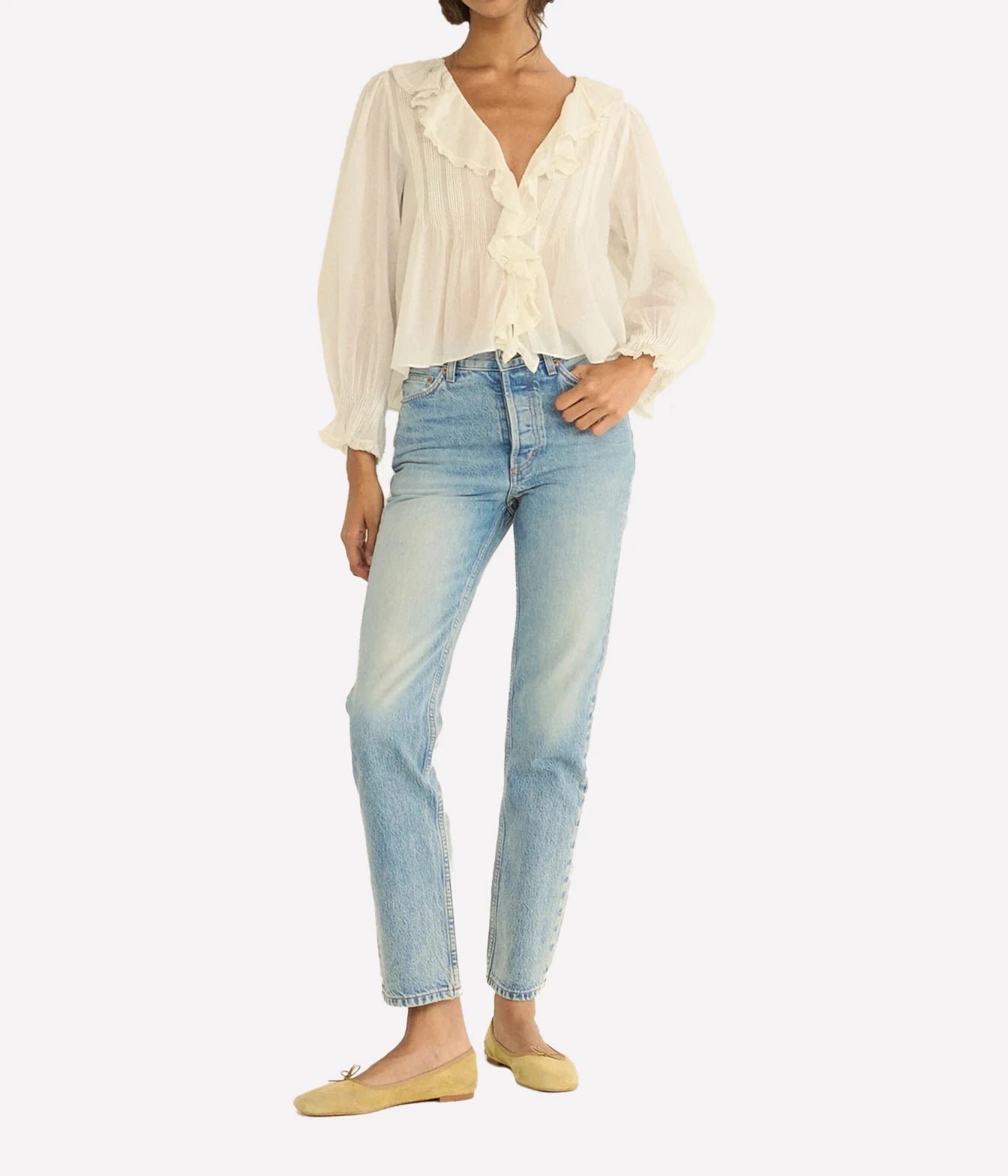 A model wearing a cream ruffled blouse, light-wash blue jeans, and beige flats, shown against a plain light gray studio background.