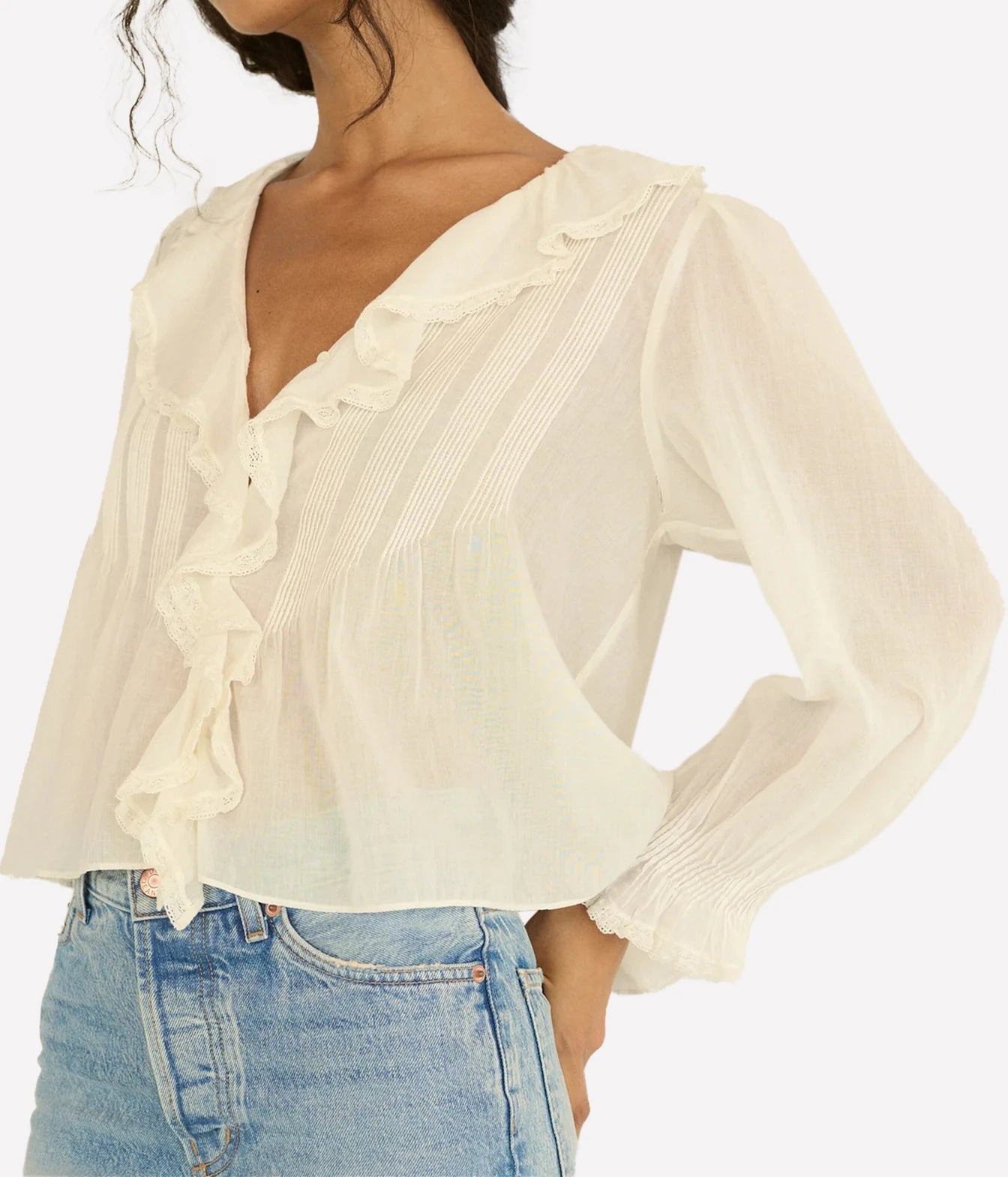 A woman wearing a cream-colored long-sleeve blouse with ruffle details and pleated texture, paired with light blue jeans, against a plain white background.