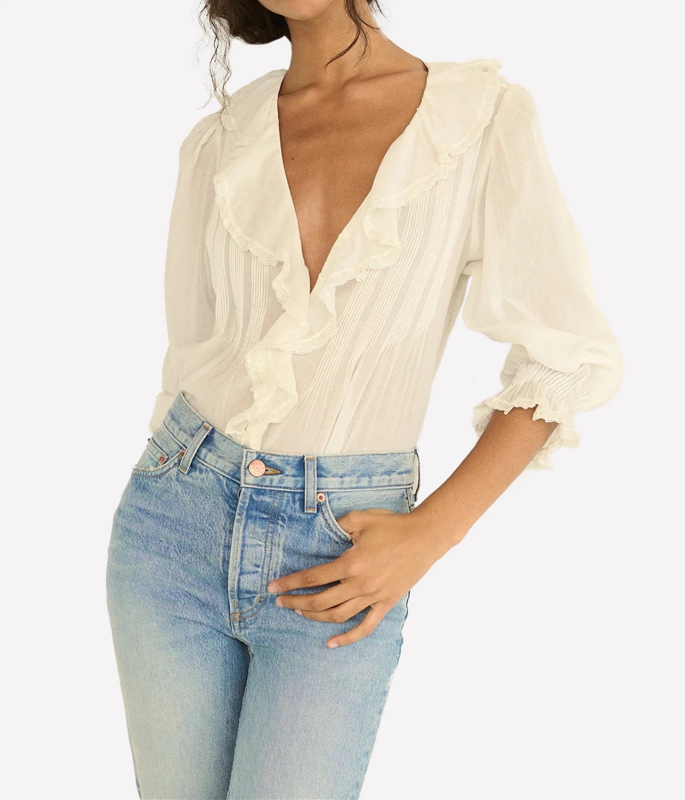 A woman wearing a cream-colored, semi-sheer blouse with dramatic ruffles and elbow-length puff sleeves, paired with light-wash blue jeans, against a plain white background.