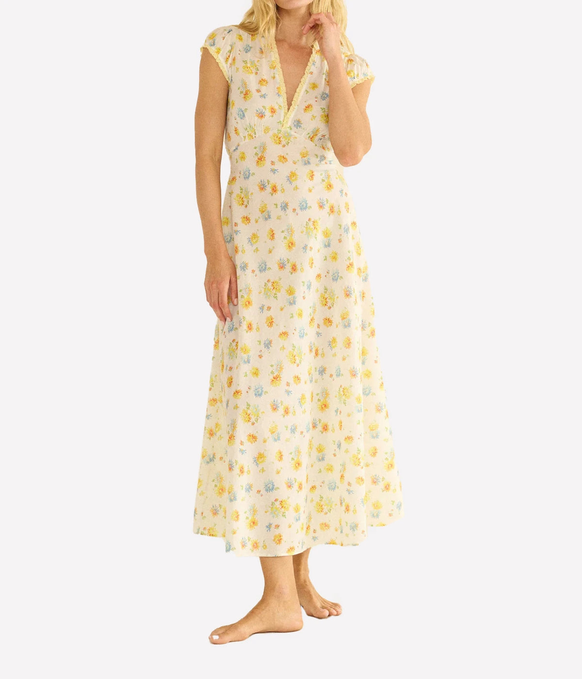 A woman modeling a sleeveless, V-neck maxi nightgown with a yellow and light blue floral pattern on a cream background, set against a plain light gray studio backdrop.
