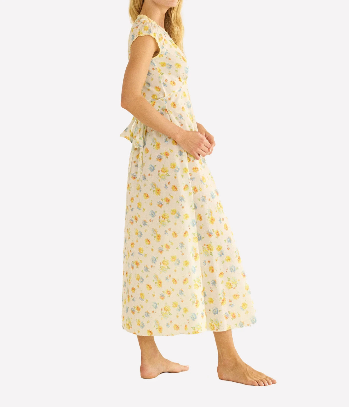 A woman modeling a sleeveless, knee-length cream dress with a yellow and light blue floral print, shown from a side profile against a plain white background.