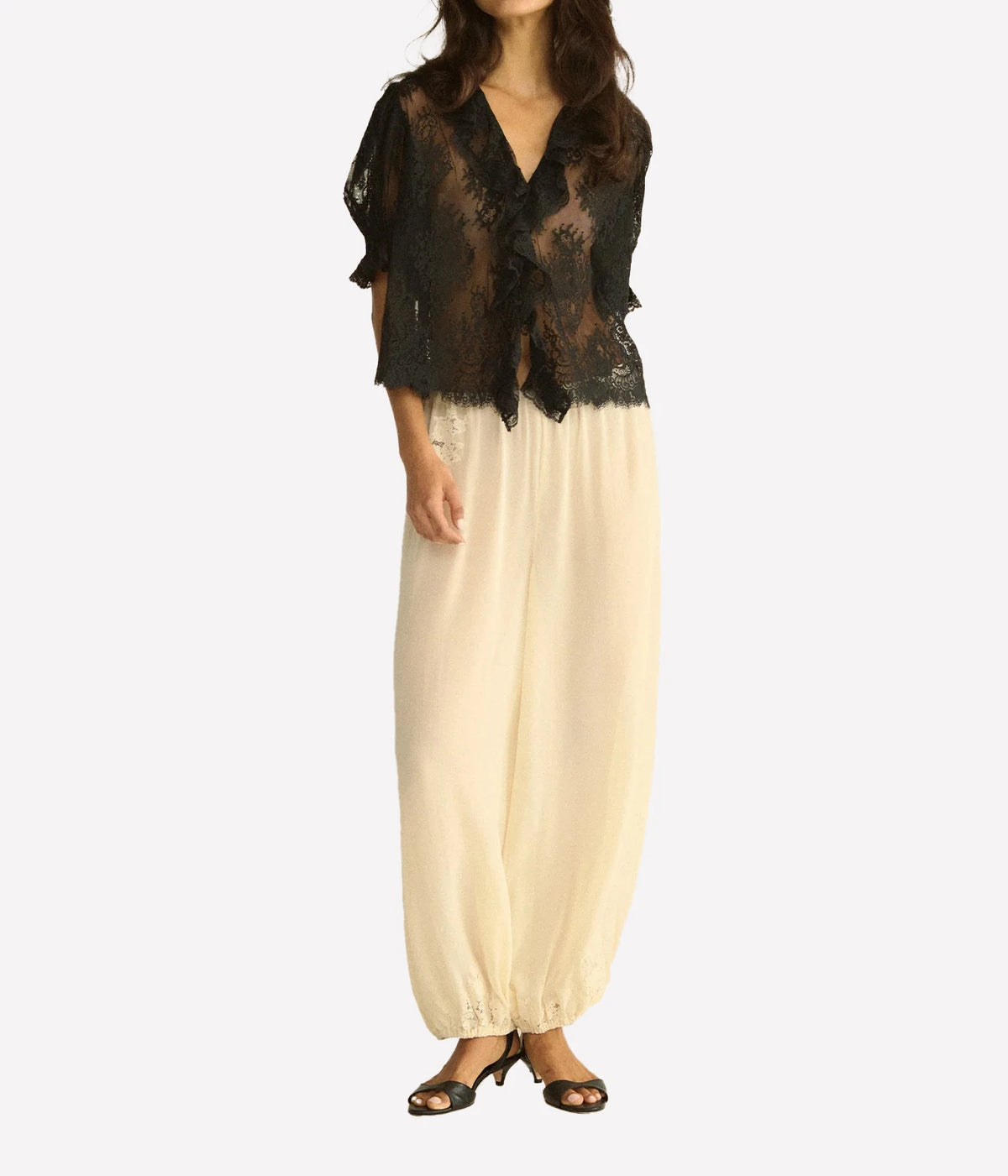 Full-length front view of a model wearing the DÔEN Lace Henri Top In Black with cream wide-leg trousers gathered at the ankle and black heeled sandals
