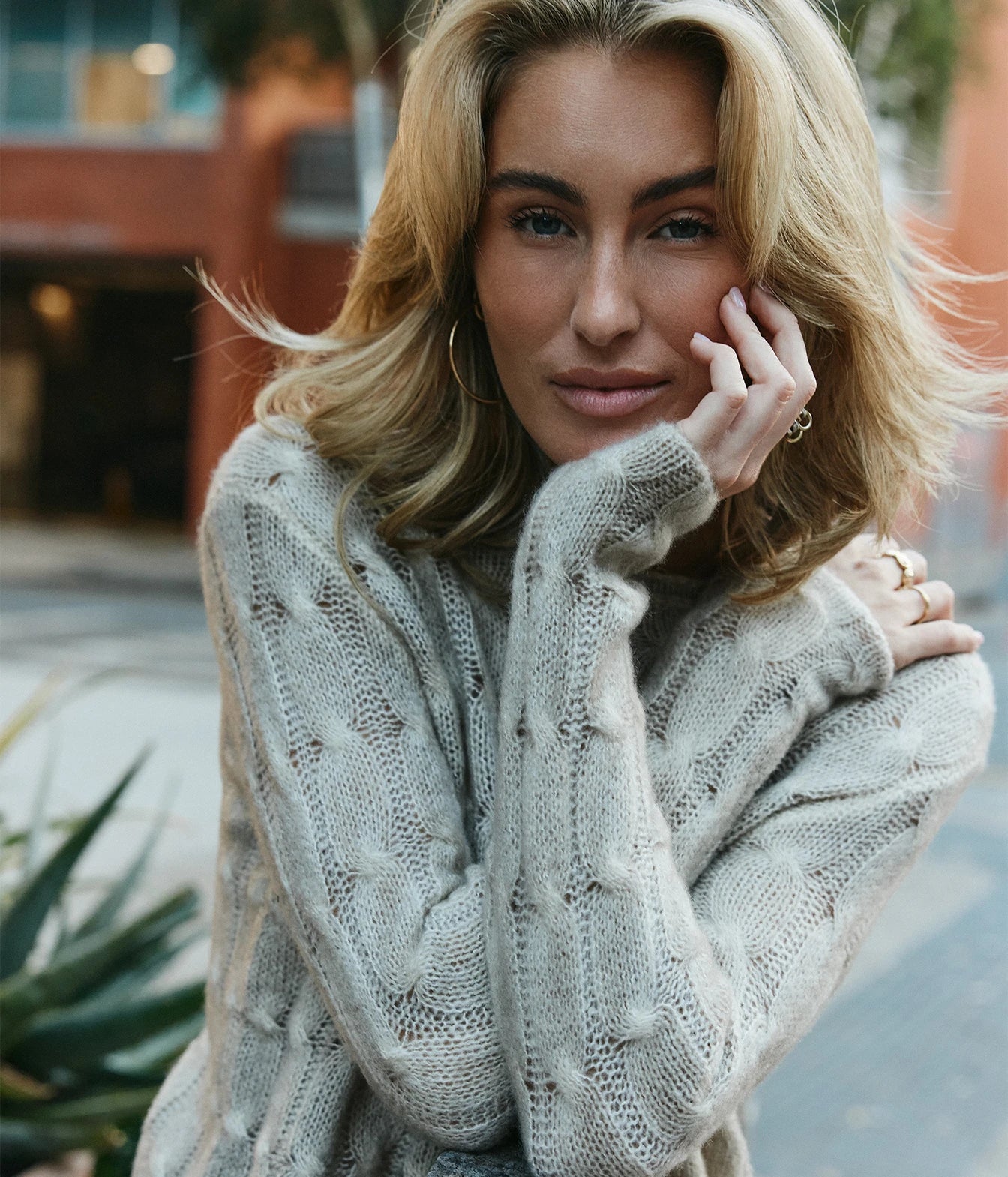 A woman wearing a light beige textured cable-knit sweater and blue jeans, standing outdoors next to a wooden bench with a blurred brick building and plants in the background.