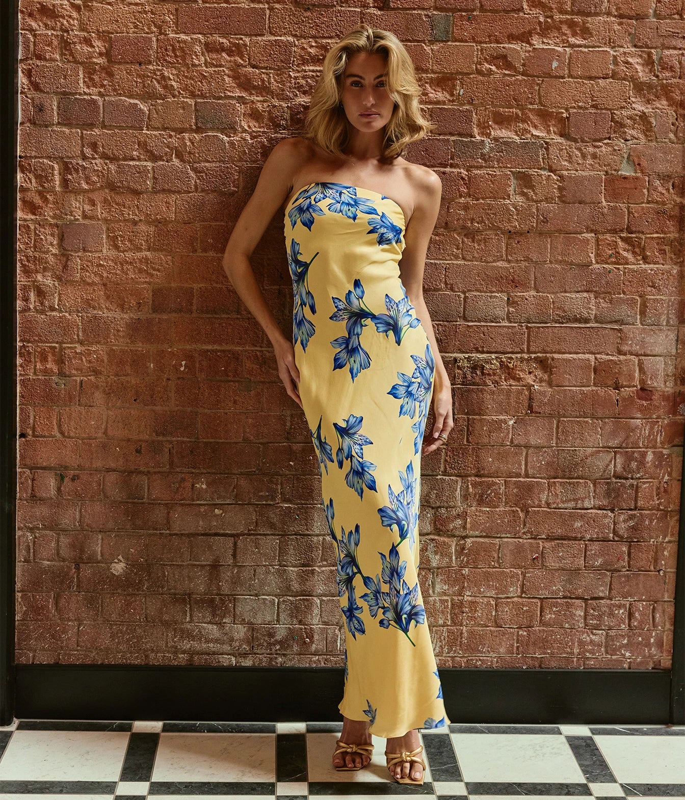A woman stands against a red brick wall wearing a strapless yellow maxi dress with a blue floral print and gold sandals.