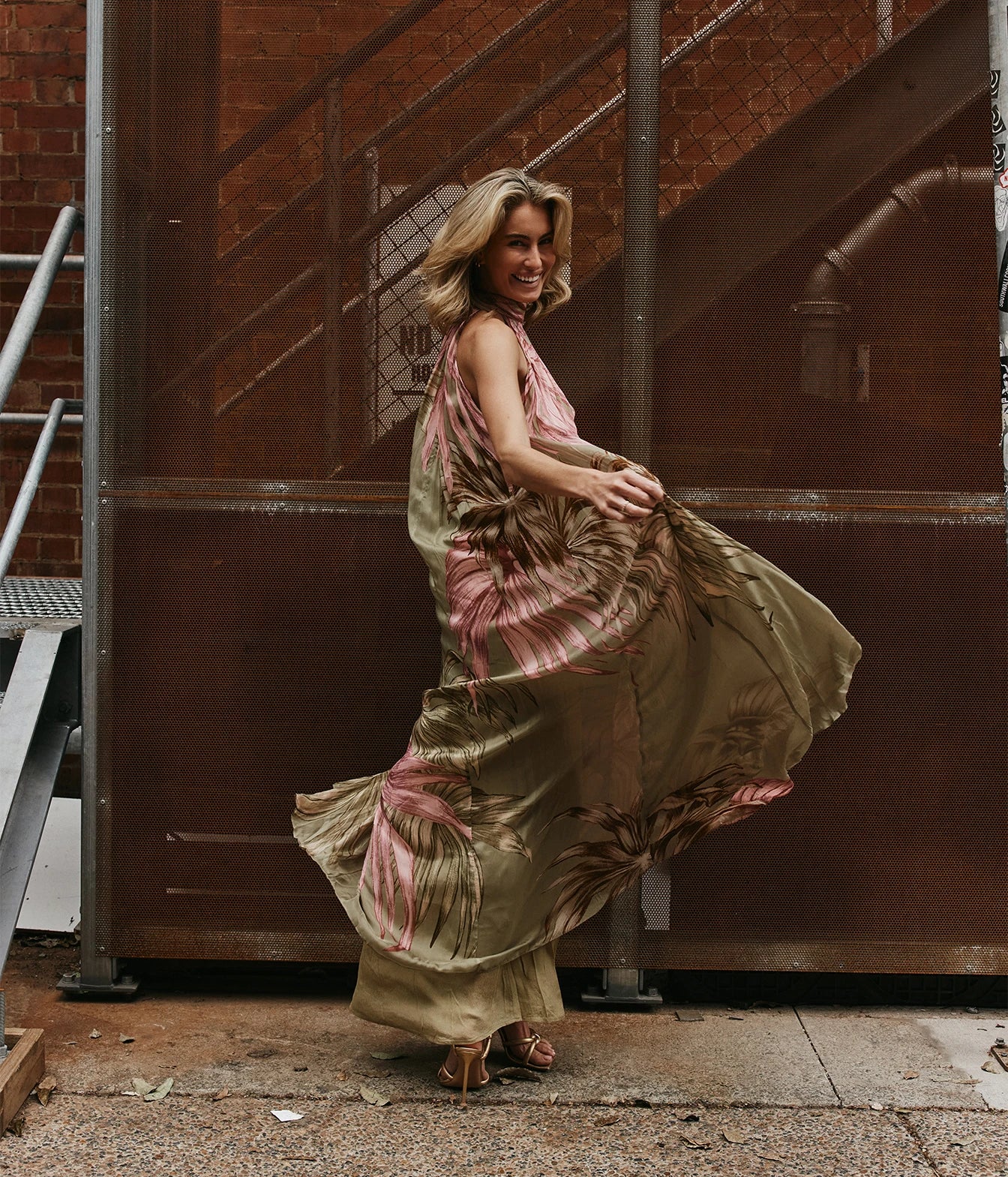 A woman wearing a flowing olive green and pink tropical-print maxi dress and brown strappy heels, captured mid-motion as she twirls in front of a large brown metal gate and brick building.