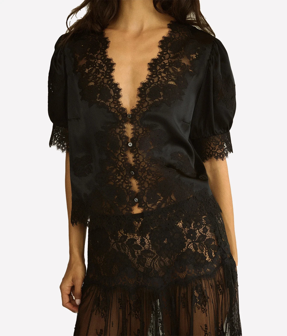 Close-up of a model wearing the DÔEN Idella Top In Black, showing the plunging scalloped lace neckline, lace-cuffed puff sleeves and the lace skirt beneath