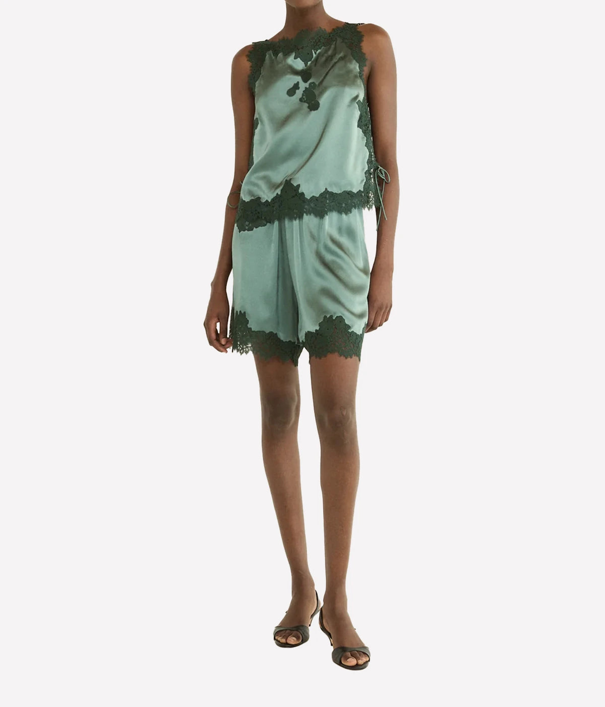 A model wearing a mint green satin romper with dark green lace trim, paired with dark brown strappy sandals, against a plain light gray background.