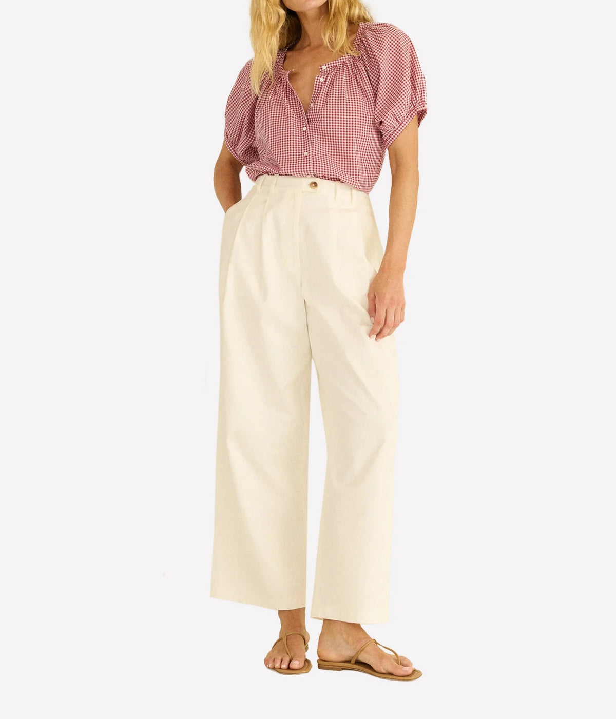 A woman modeling a pink and white gingham short-sleeve blouse tucked into high-waisted, wide-leg cream trousers, paired with tan sandals, against a plain white background.