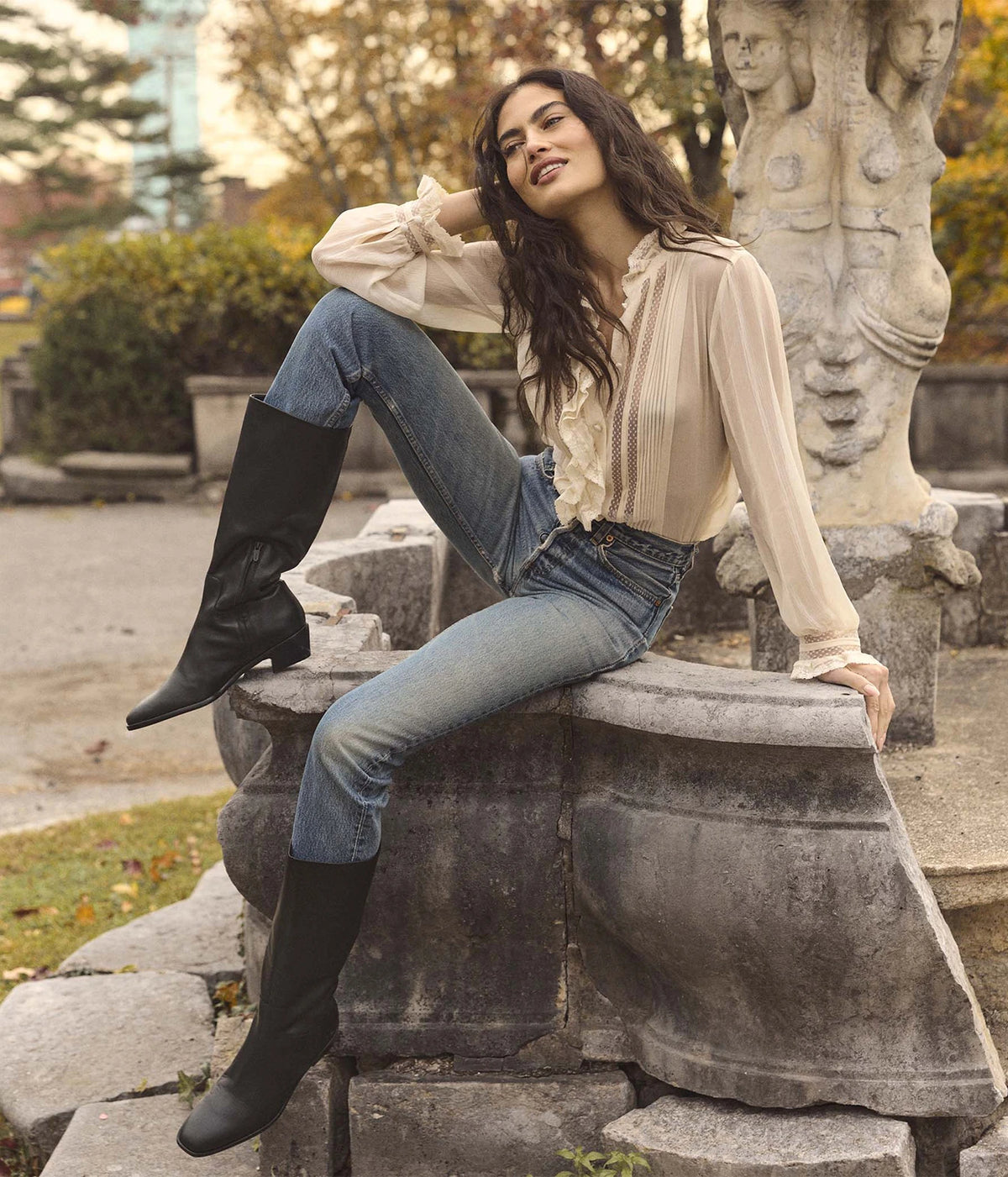 Model seated wearing the DÔEN Magdeleine Top In Candlelight with light-wash jeans and black knee-high boots, one knee raised