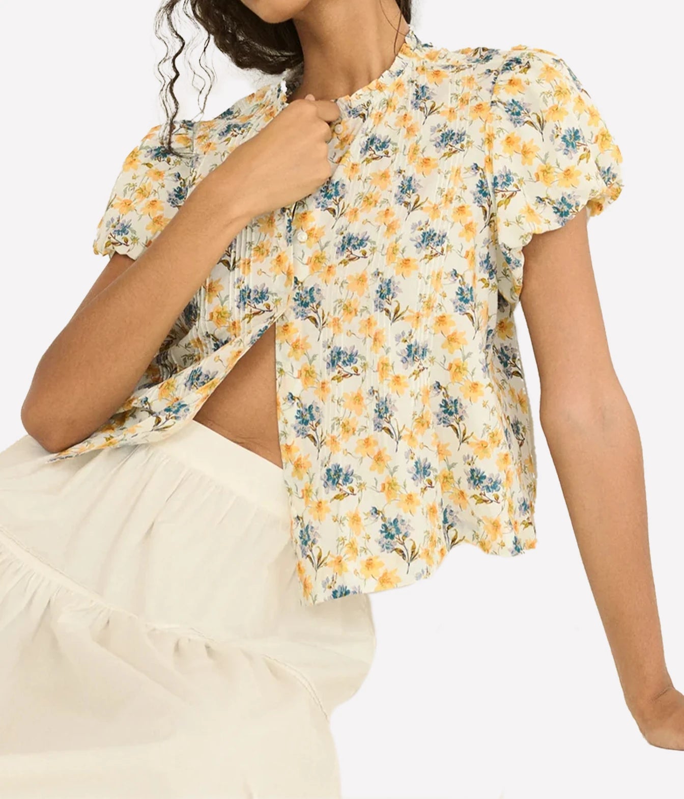A woman wearing a short-sleeve floral blouse with puff sleeves, a round neckline, and a side slit, paired with an off-white skirt. The blouse features a yellow and blue floral pattern on a cream background.