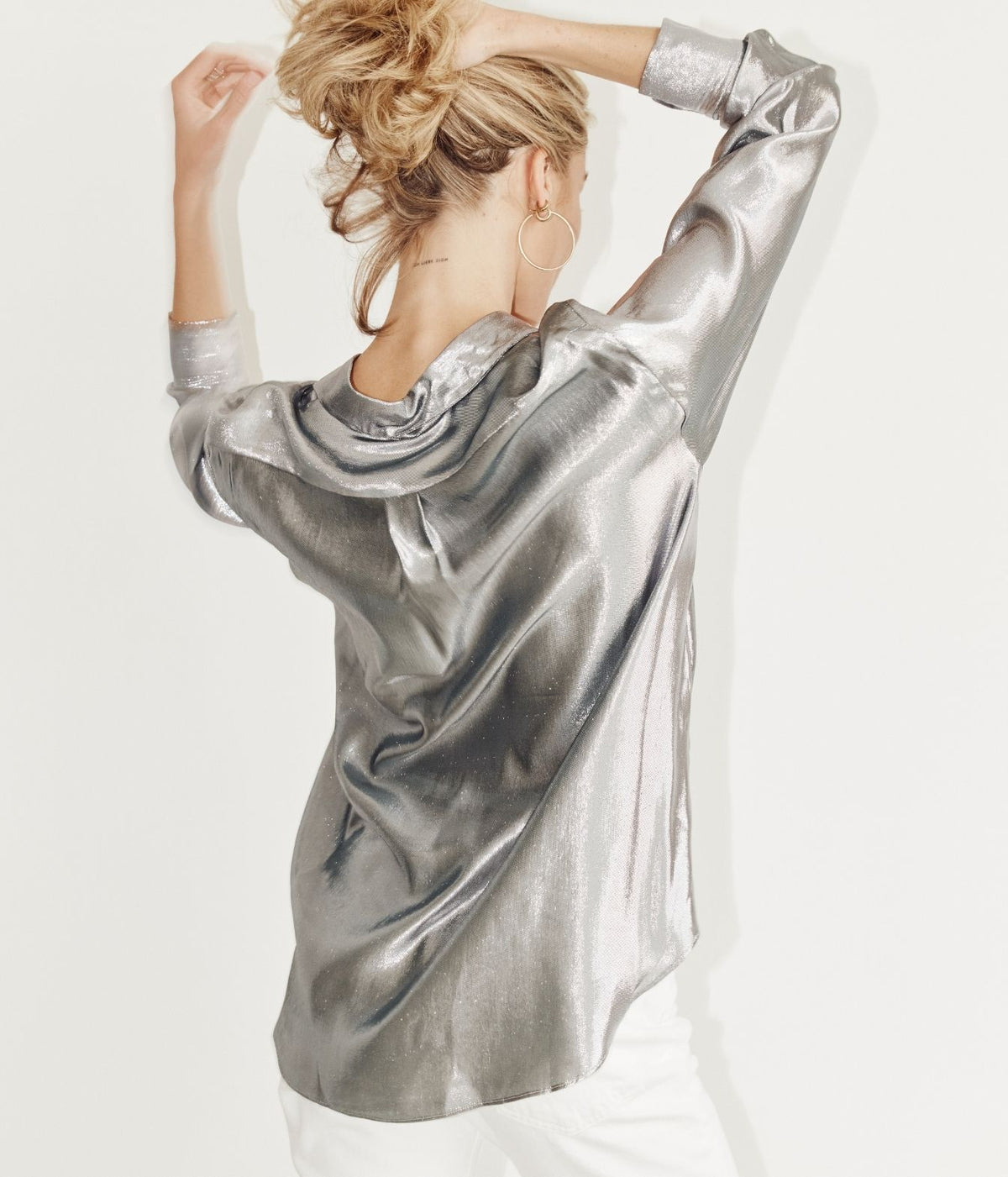 A woman with blonde hair in a messy bun is shown from behind wearing a shiny silver metallic long-sleeve top and white pants against a plain white background.