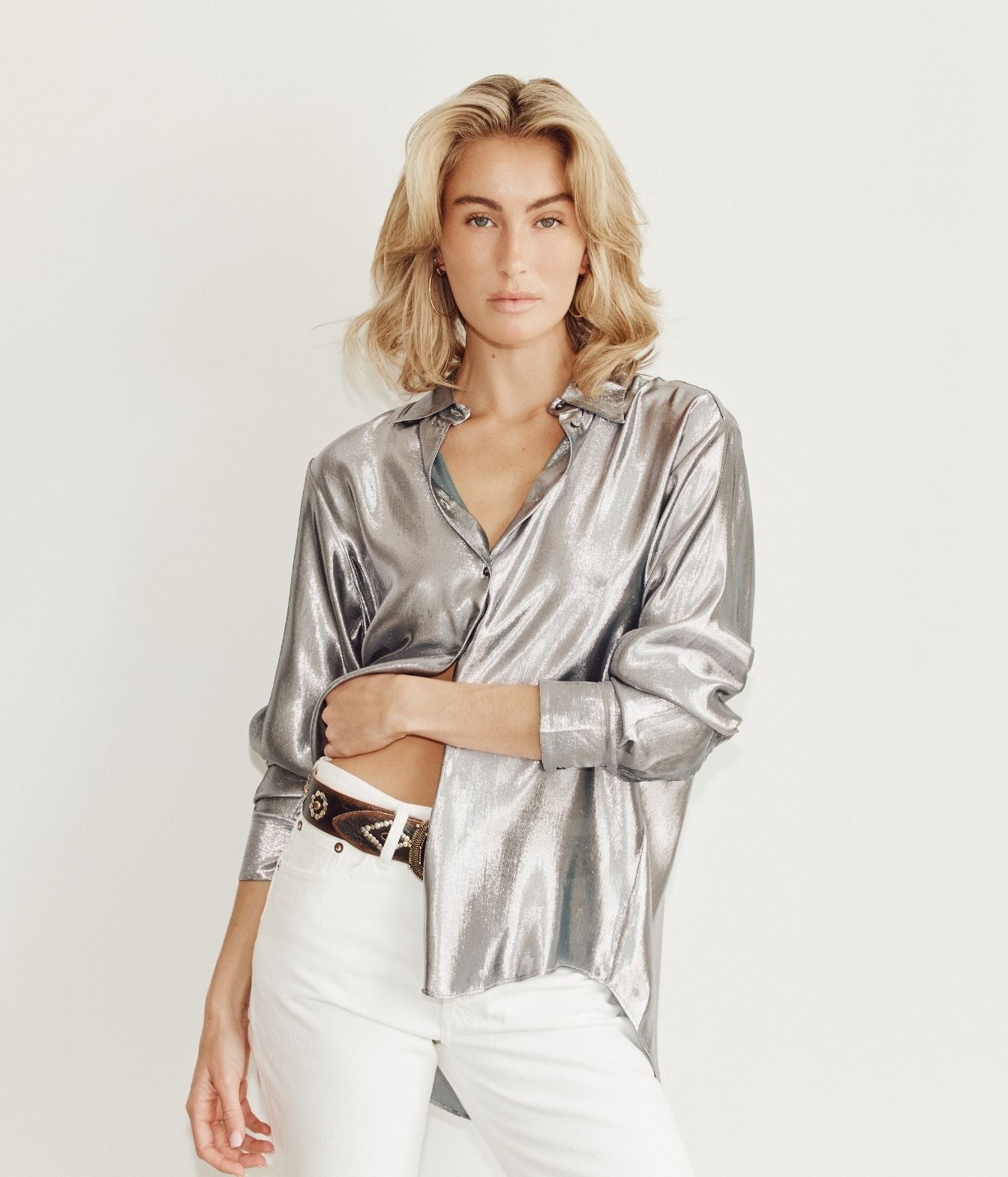 A woman modeling a shiny metallic silver button-up shirt with white pants and a brown belt, posed against a clean white studio background.