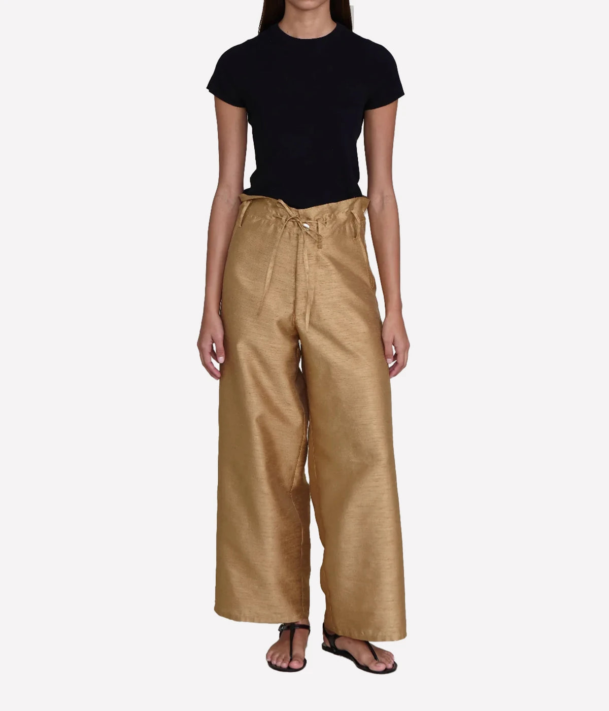 A model wearing a black short-sleeve crewneck top and high-waisted, wide-leg metallic gold trousers with a drawstring waist, paired with black sandals, against a light gray background.