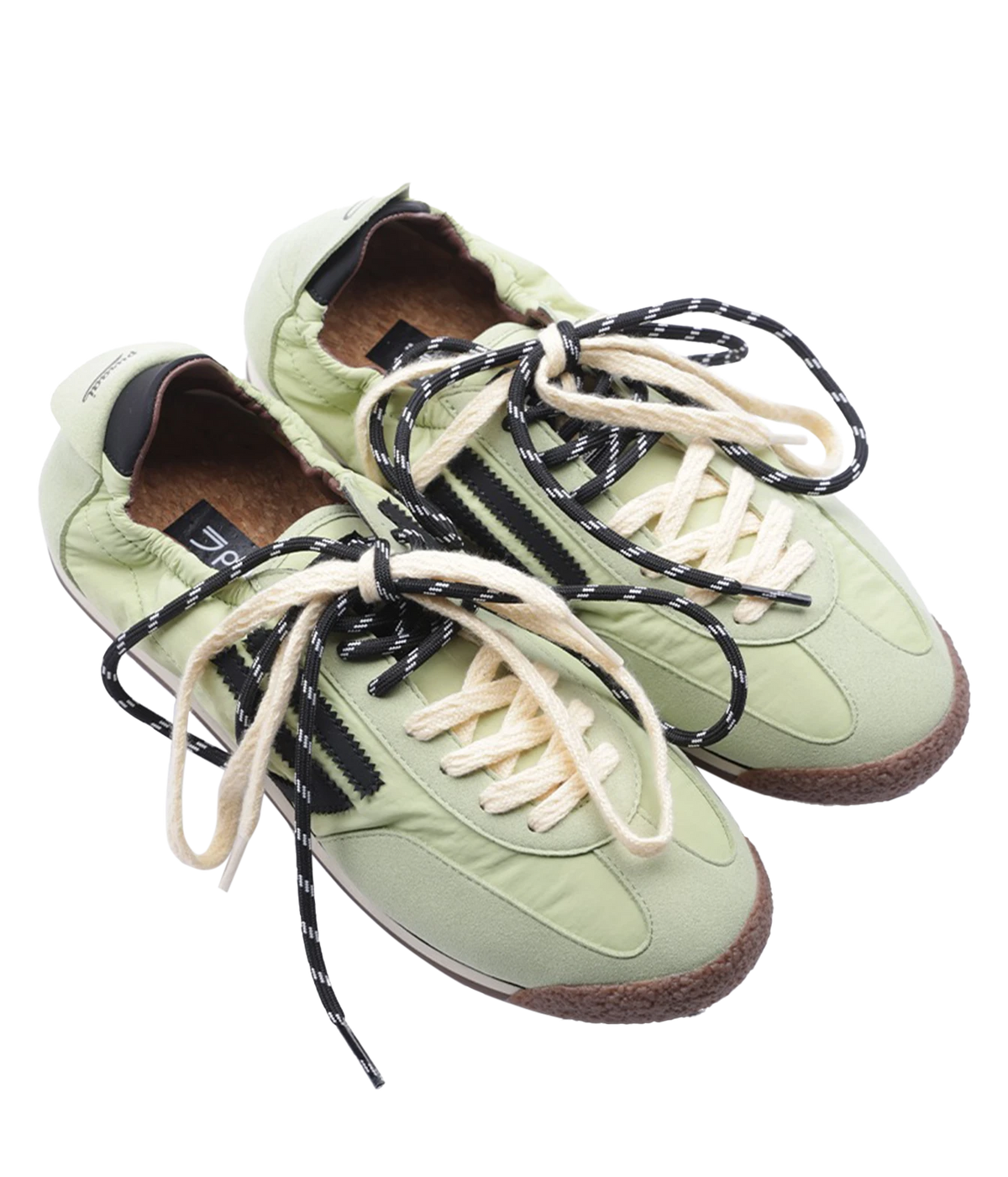 A pair of light sage green sneakers with black and white patterned laces, black side stripes, and brown midsoles, shown on a plain white background.