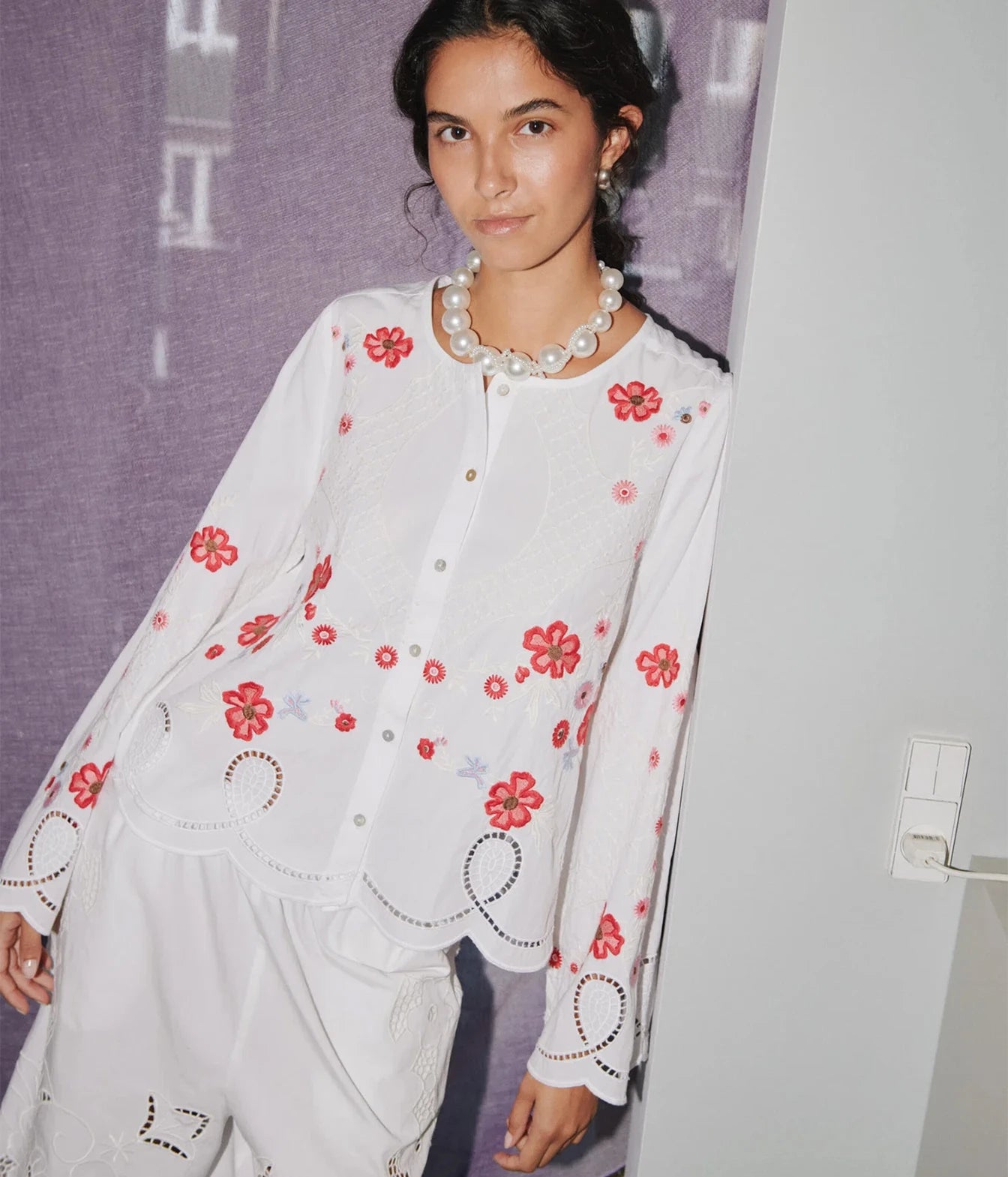A woman wearing a white long-sleeve blouse with red floral embroidery and scalloped edges, paired with white embroidered pants, standing against a purple textured backdrop.