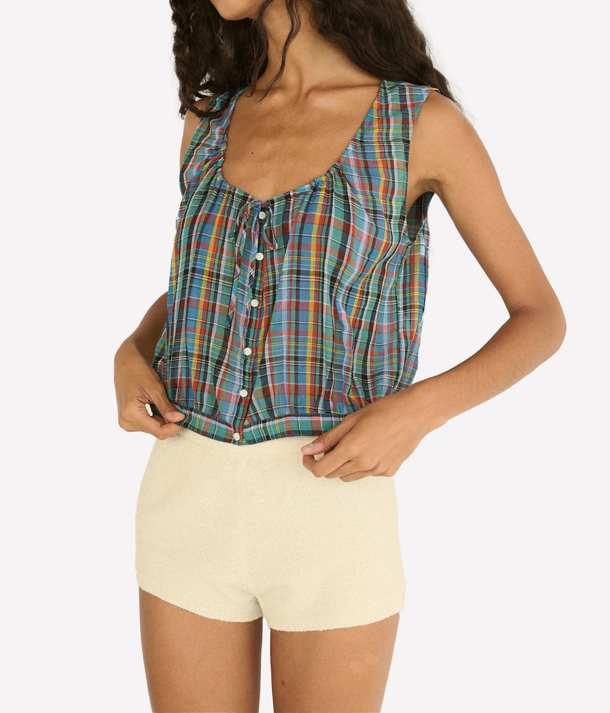 A woman wearing a sleeveless plaid crop top with a front button placket and tie detail, paired with cream shorts, against a light gray studio background.