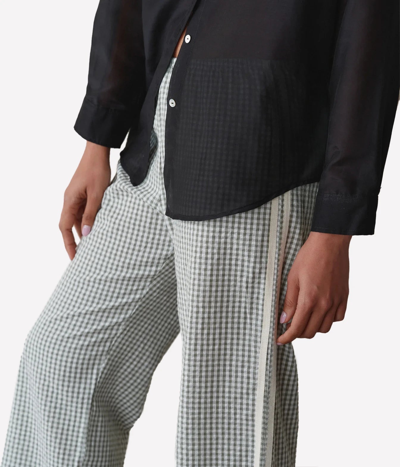 A close-up of a person wearing wide-leg trousers in a black and white gingham pattern with a white side stripe, paired with a black button-up shirt, against a light gray background.