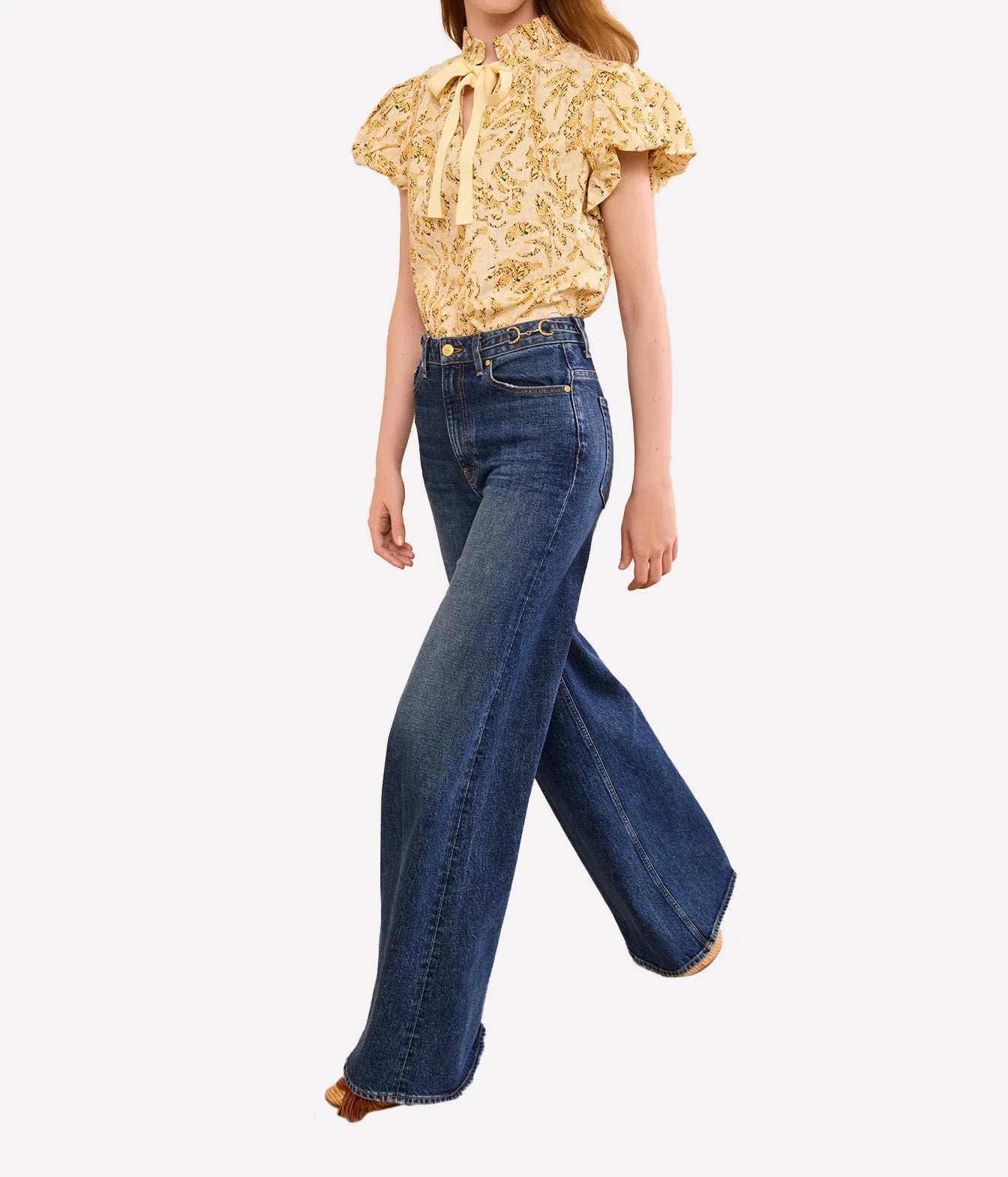 A model wearing a yellow and white patterned ruffled blouse with a tie-neck and dark blue wide-leg denim jeans, shown from the waist down against a light gray background.