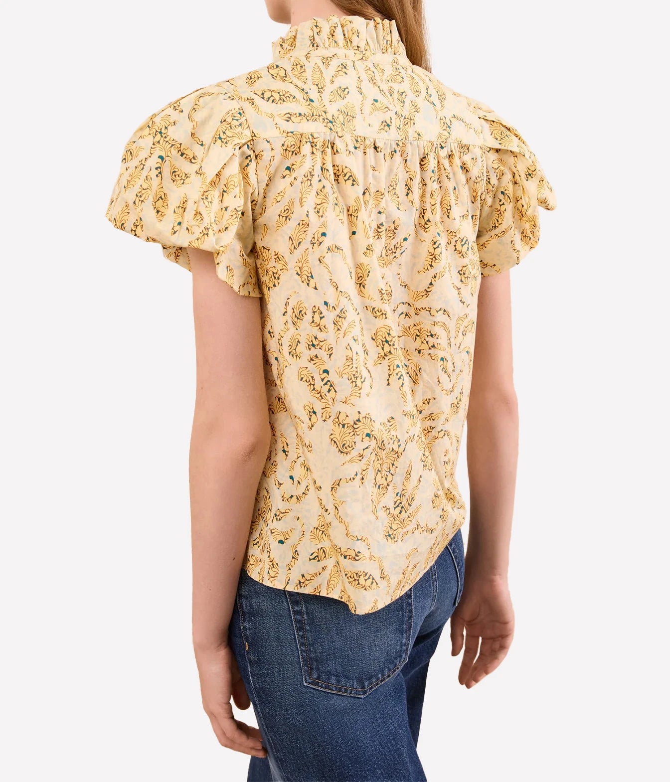 A woman wearing a cream-colored blouse with a yellow and green paisley pattern, featuring ruffled short sleeves and a smocked neck, paired with blue jeans against a light gray background.