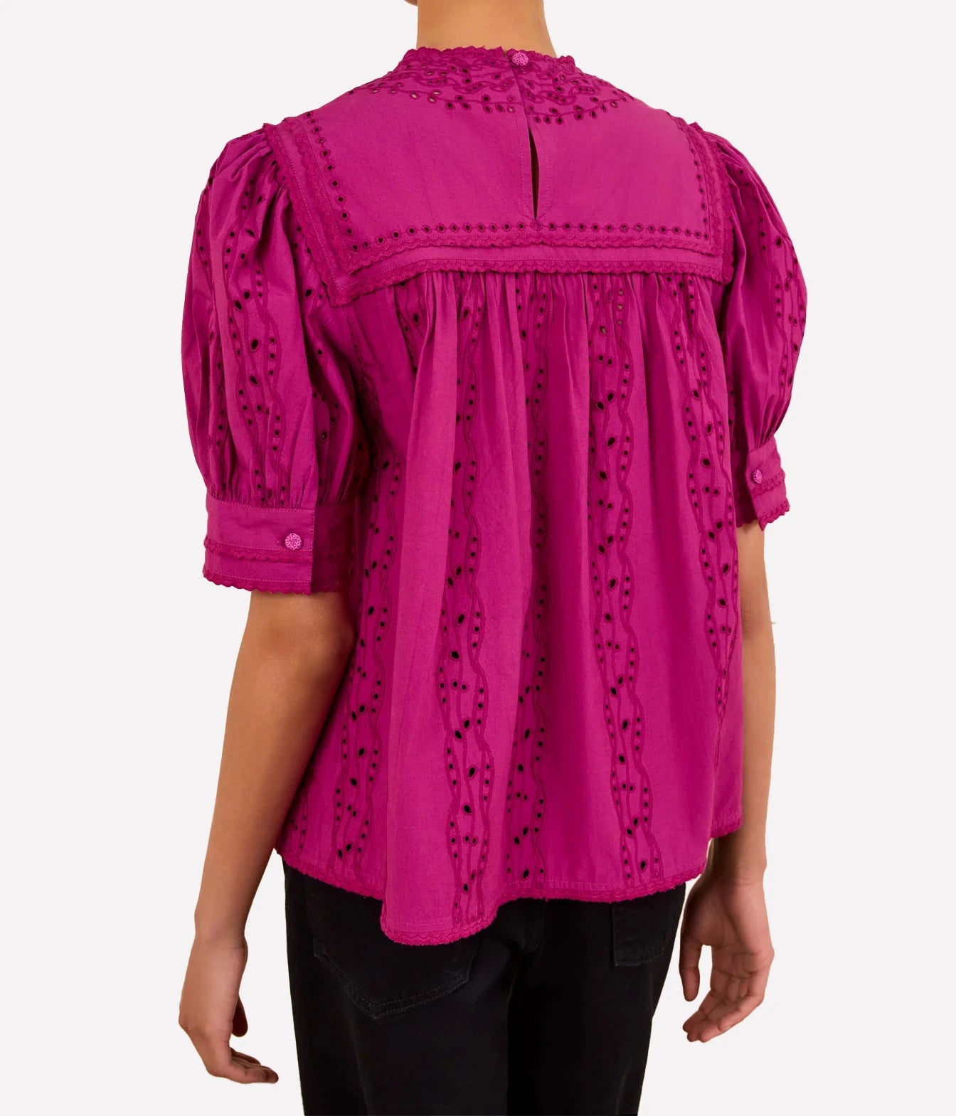 A woman wearing a bright magenta eyelet blouse with puff sleeves and a high neckline, shown from the back against a light gray studio background.