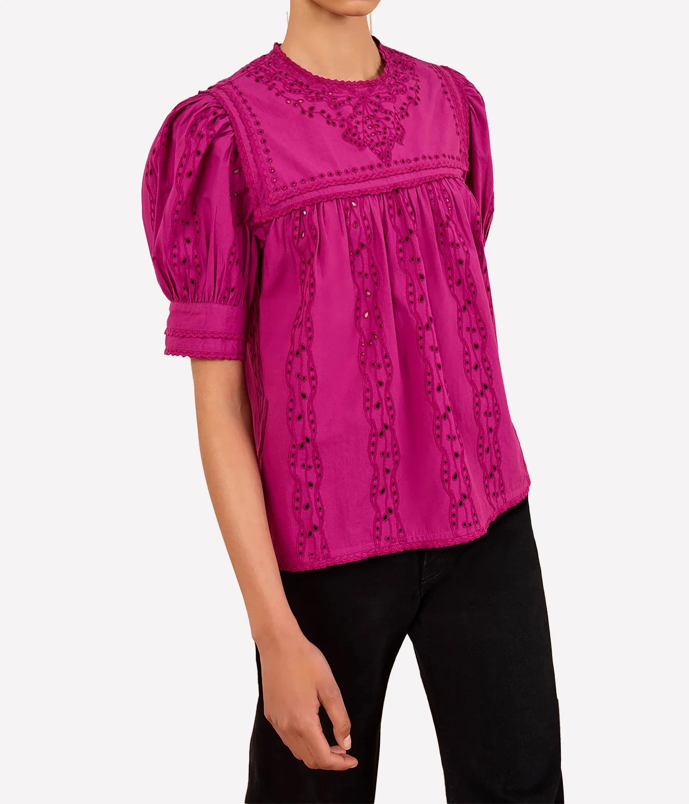 A woman wearing a bright magenta pink eyelet blouse with puff sleeves and black pants, shown from the neck down against a light gray background.