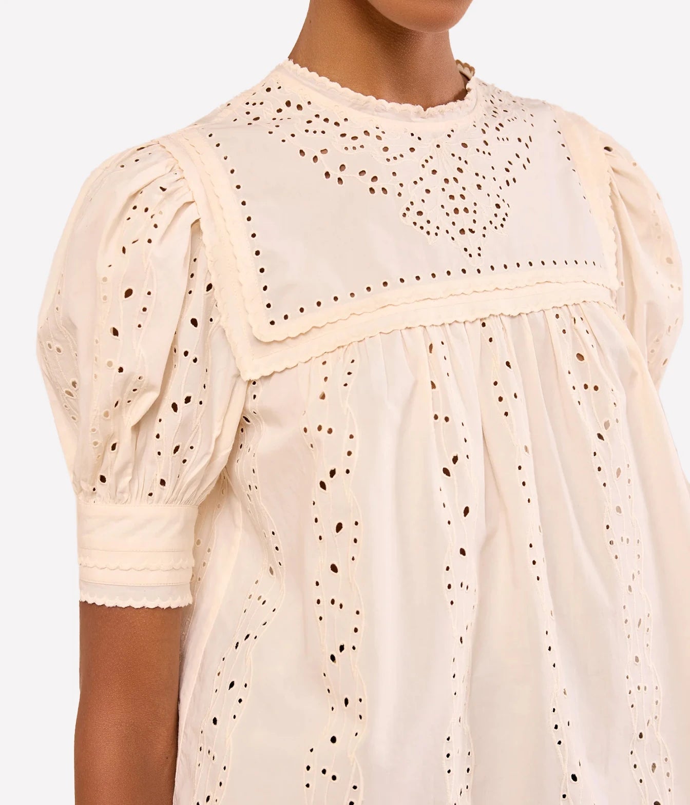 A close-up of a cream-colored eyelet cotton dress with puffed short sleeves and a scalloped neckline, shown on a model against a plain white background.
