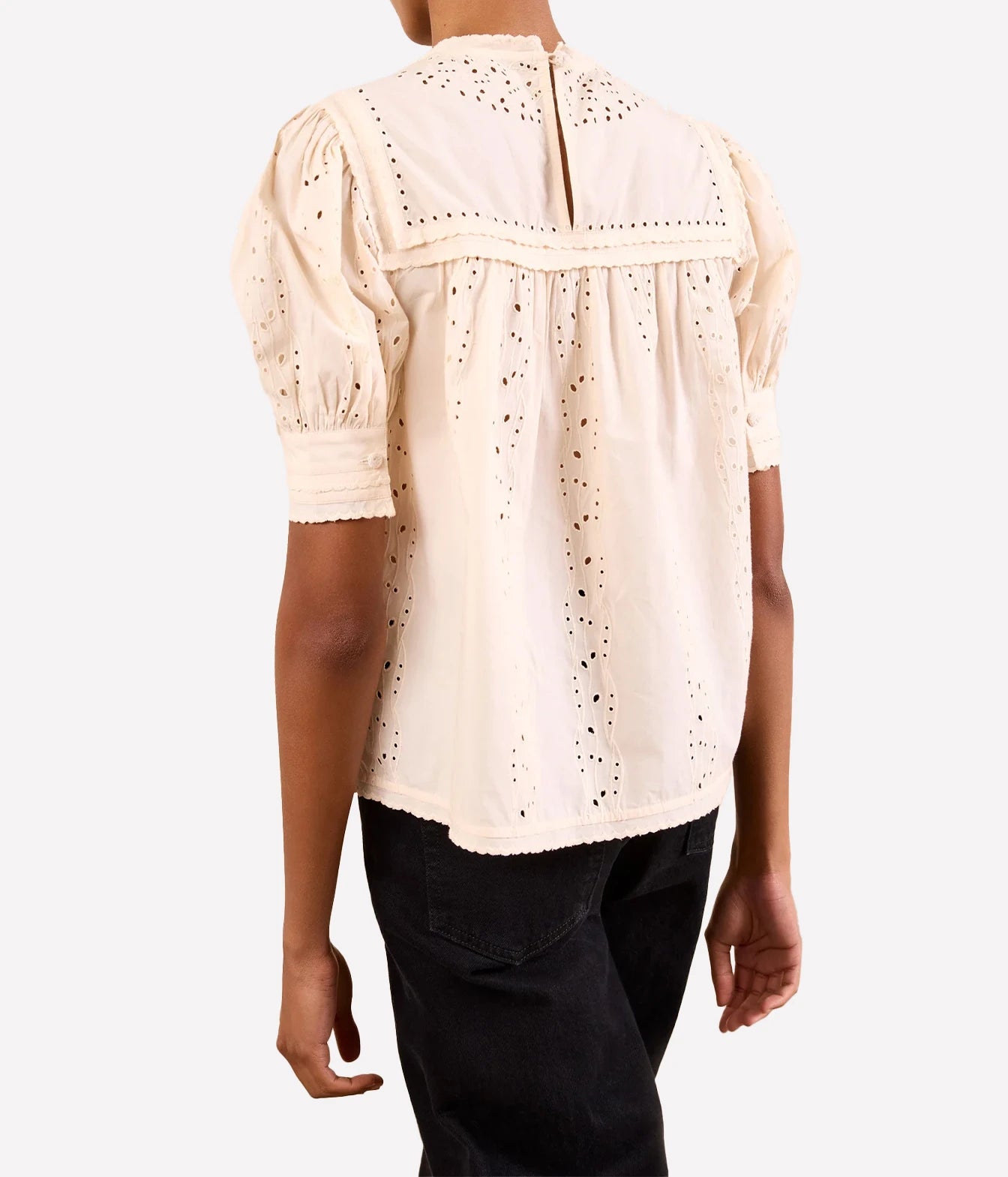 A person shown from the back wearing a cream-colored eyelet blouse with puff sleeves and black pants, photographed against a light gray studio background.