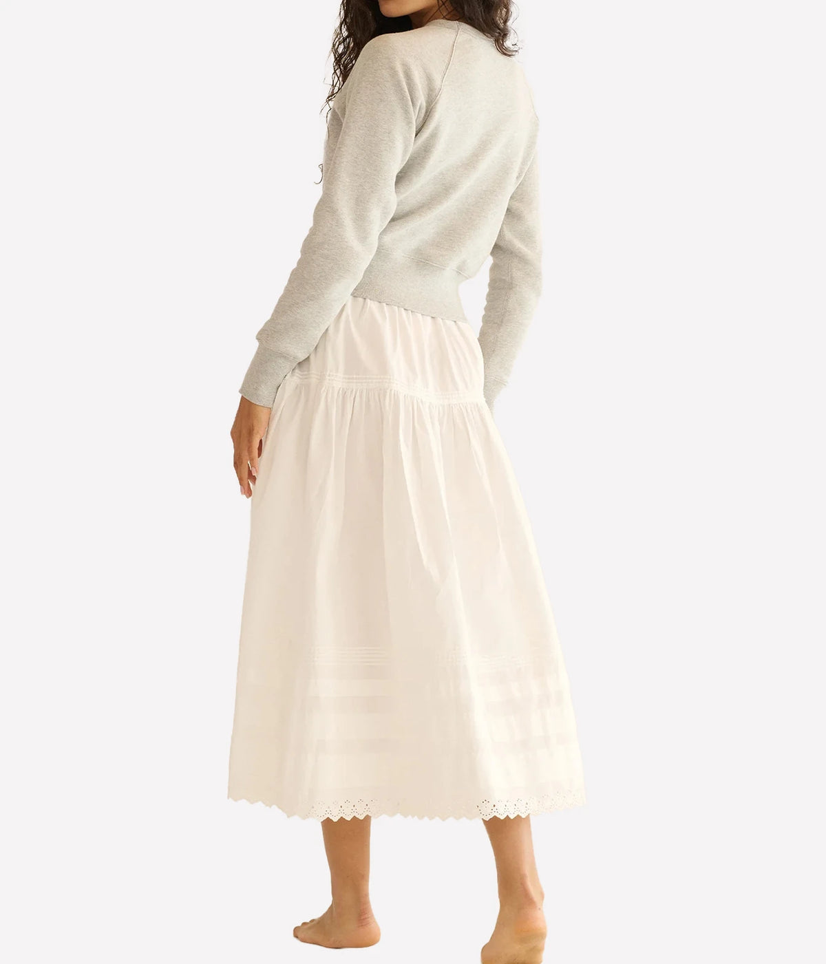 A woman shown from the back wearing a light beige knit sweater and a cream-colored midi skirt with a scalloped hem, against a plain light gray studio background.