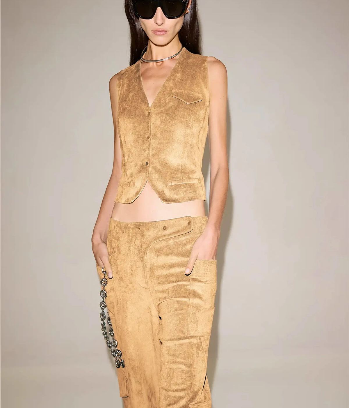 Front view of a model wearing the Alexis Aquila Vest In Dune buttoned with a V-neckline, chest welt pocket and cropped hem, styled with matching suede-look cargo trousers, black sunglasses, a silver choker and a silver hanging chain at the hip