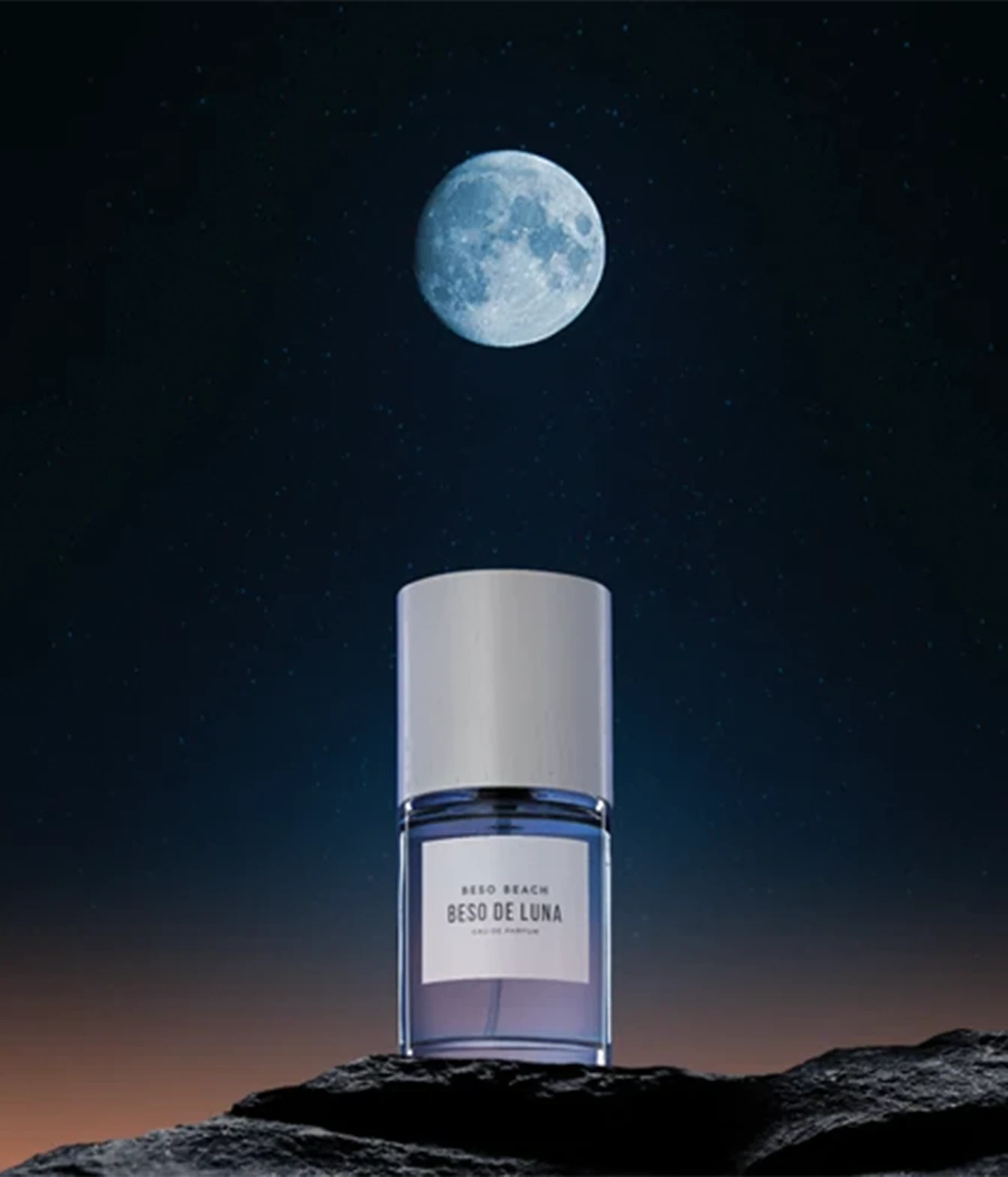 A bottle of Aedo Beach Besi de Luna body spray stands on a dark rocky surface against a night sky with a large moon and stars. The bottle is clear with a lavender-purple gradient liquid and a white cap.