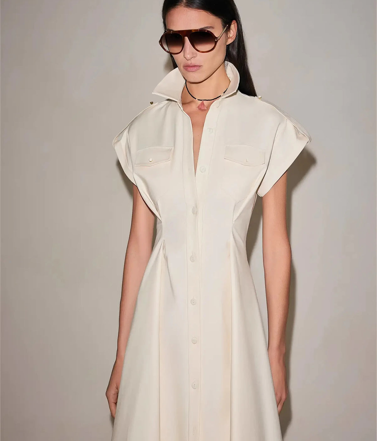 Three-quarter front view of a model wearing the Alexis Ceres Dress In Beige with brown sunglasses, showing the cap sleeves, buttoned bodice and flared skirt