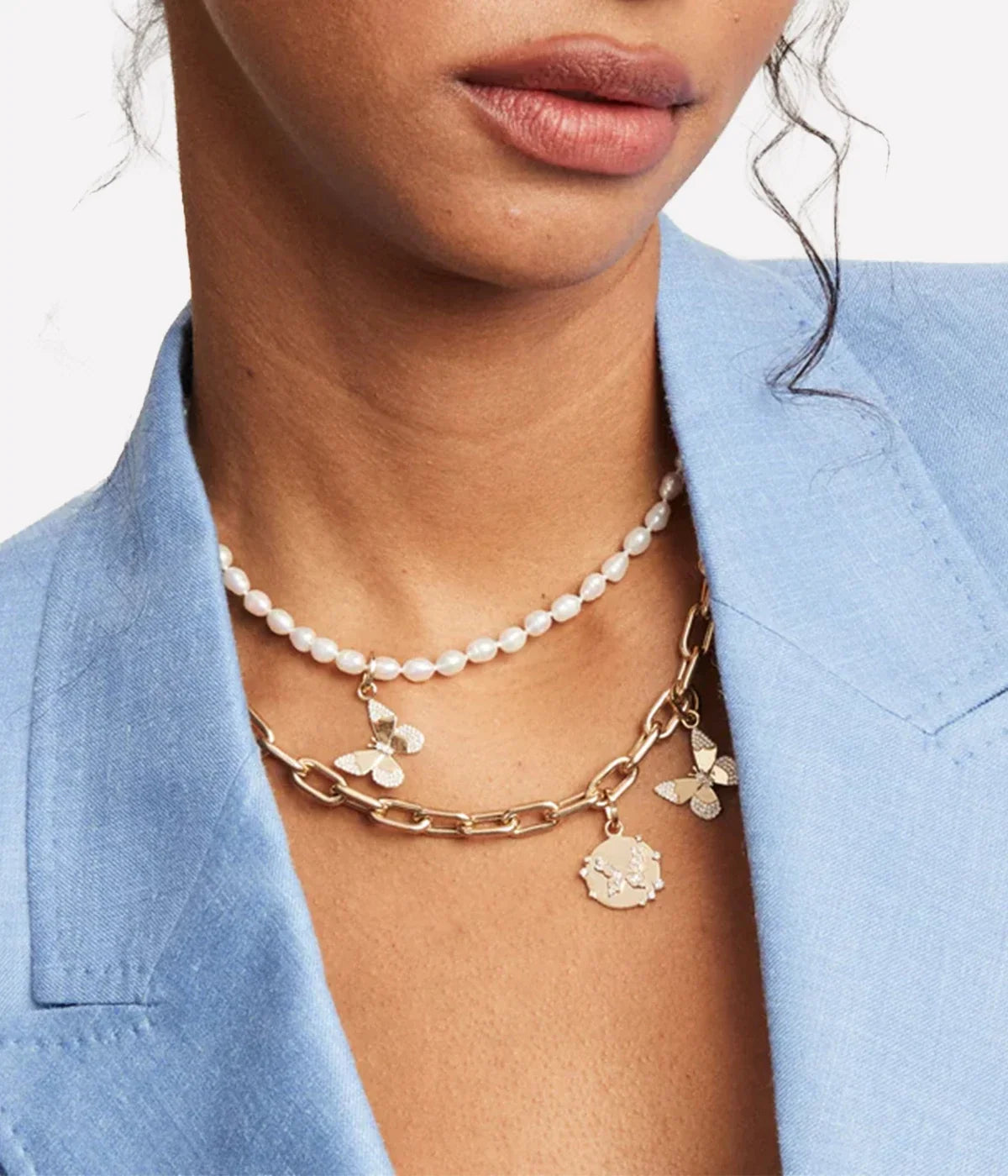 Close-up of a model wearing the Adina Reyter Diamond Butterfly Charm in 14k Yellow Gold on a gold chain alongside a round gold charm, layered under a pearl necklace with a light blue blazer