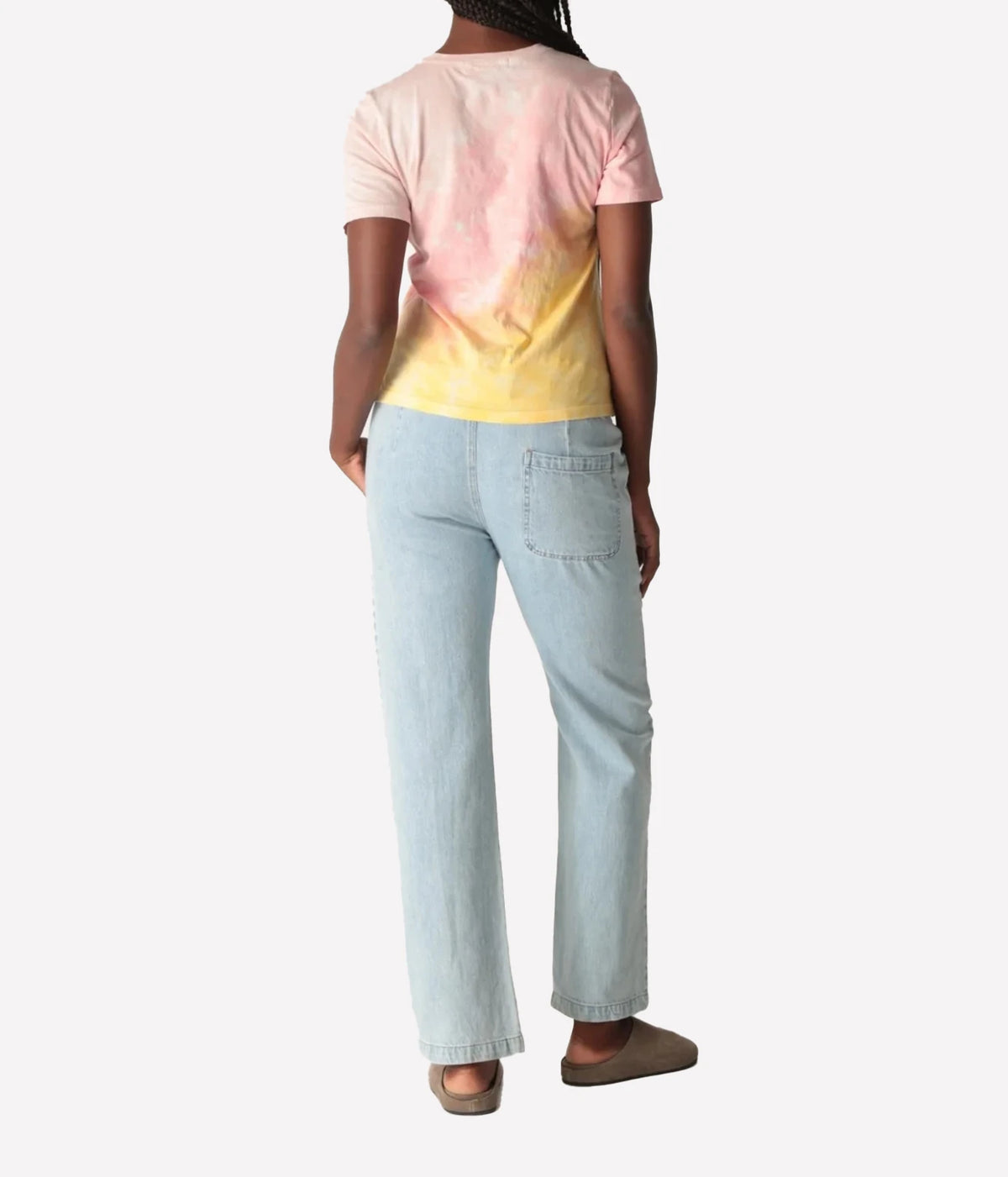 Back view of a model wearing the Electric Rose Everyday Haze T-Shirt in Sunrise Multi, showing the pink-to-yellow fade across the back, with light-wash straight-leg denim pants and taupe suede clogs
