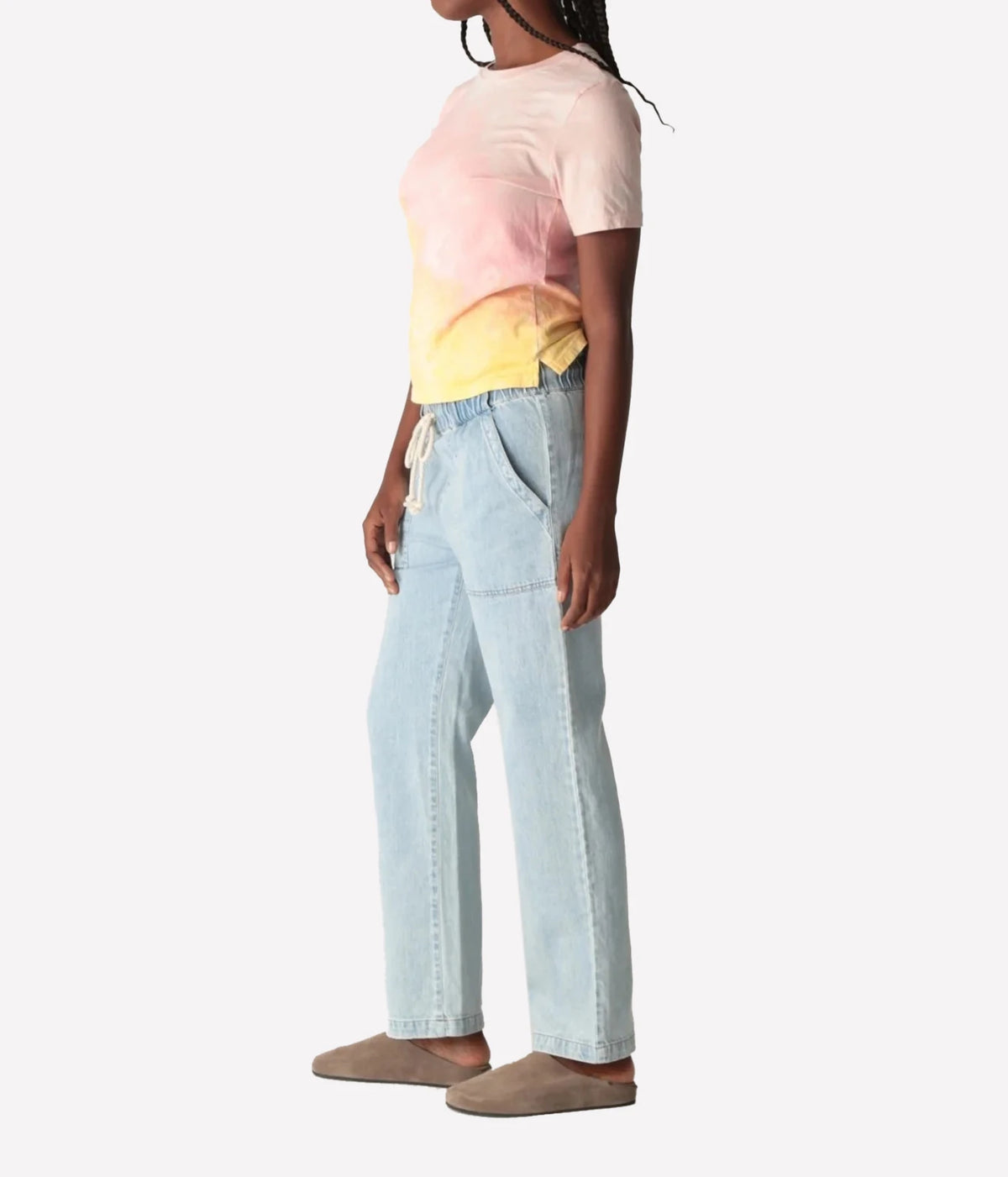 Side profile of a model wearing the Electric Rose Everyday Haze T-Shirt in Sunrise Multi, showing the cropped boxy fit and side splits, with light-wash straight-leg denim pants and taupe suede clogs