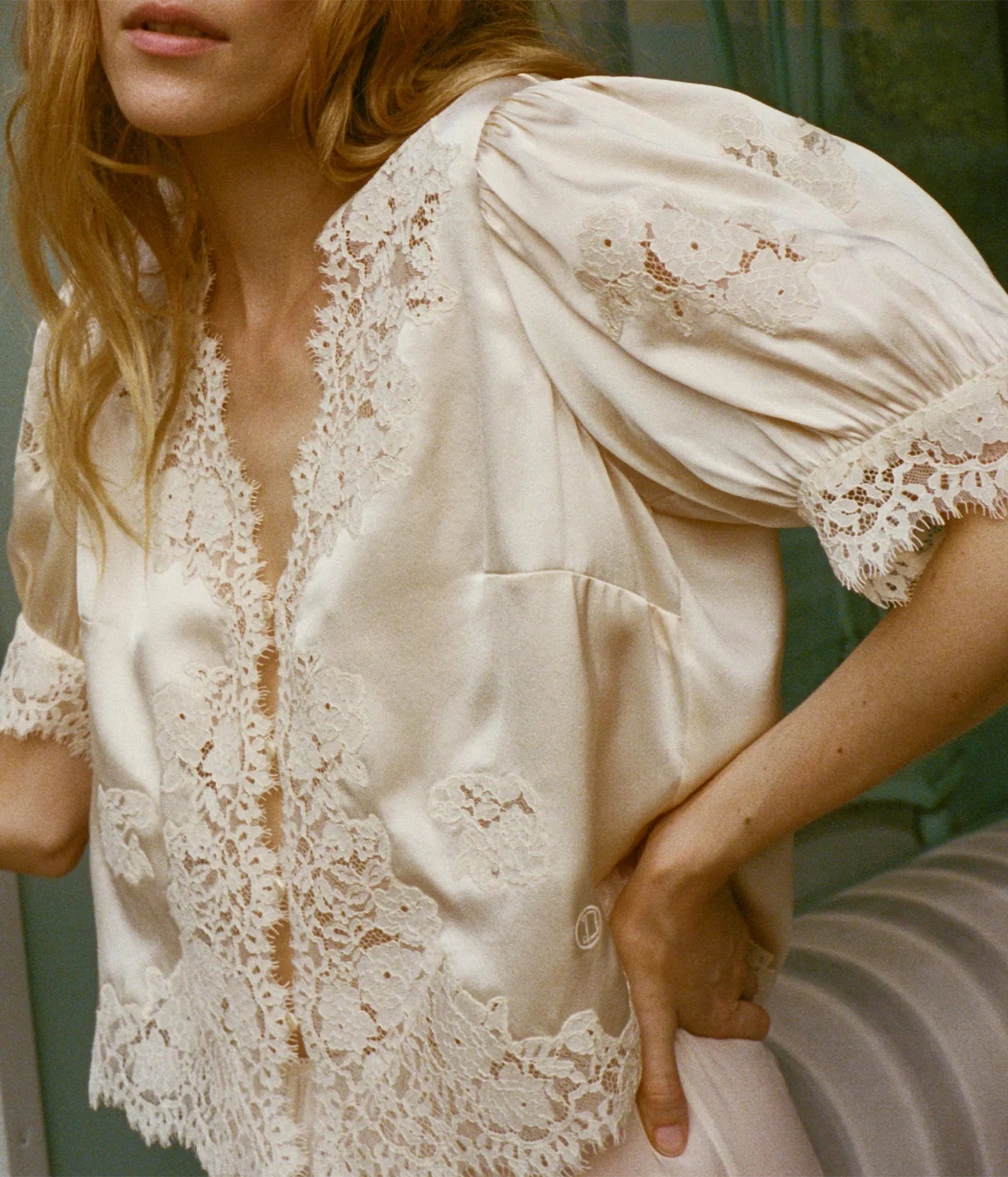 A close-up of a woman wearing a cream satin blouse with intricate floral lace trim along the neckline, front placket, pockets, and cuffs.