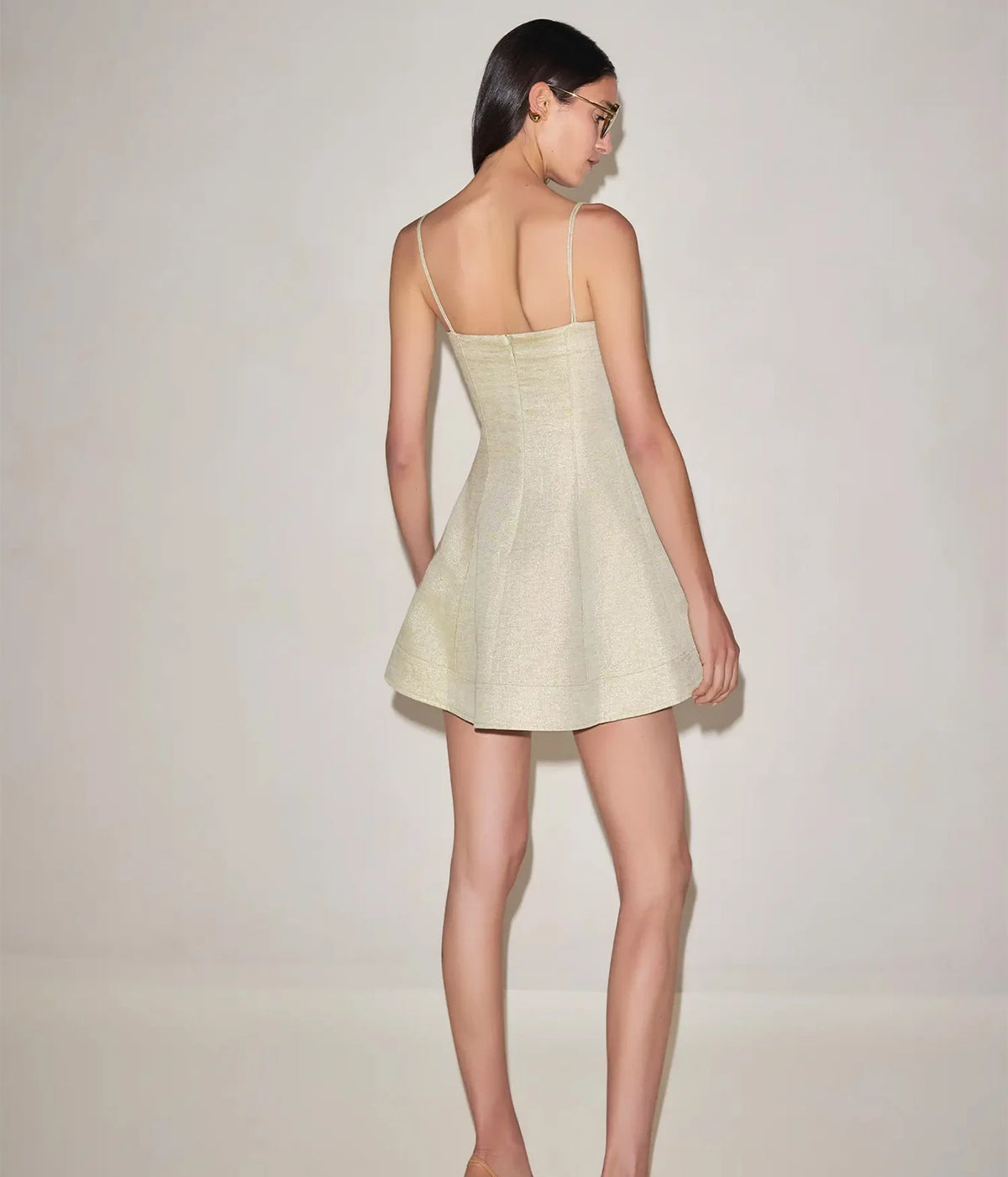 Full-length back view of a model wearing the Alexis Josefin Dress In Gold Mist with her face turned over her shoulder, showing the braided straps, fitted back and flared mini skirt
