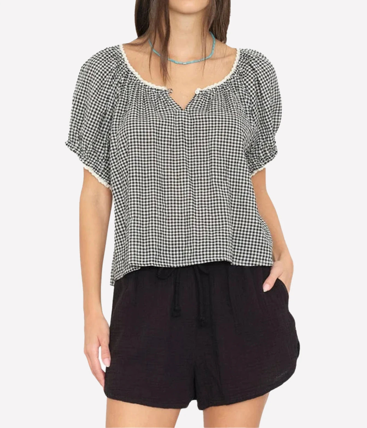 Model wearing the Xirena Maisie Top in Black Sand loose over black drawstring shorts with a turquoise beaded necklace, showing the notched neckline, contrast trim and puff sleeves