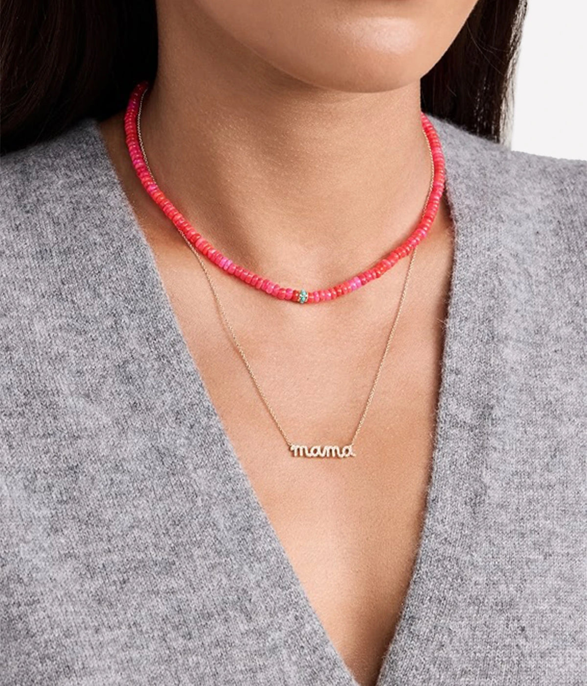 Model wearing the Sydney Evan Mama Script Necklace In 14K Yellow Gold on a fine chain layered under a bright coral beaded necklace with a grey V-neck knit