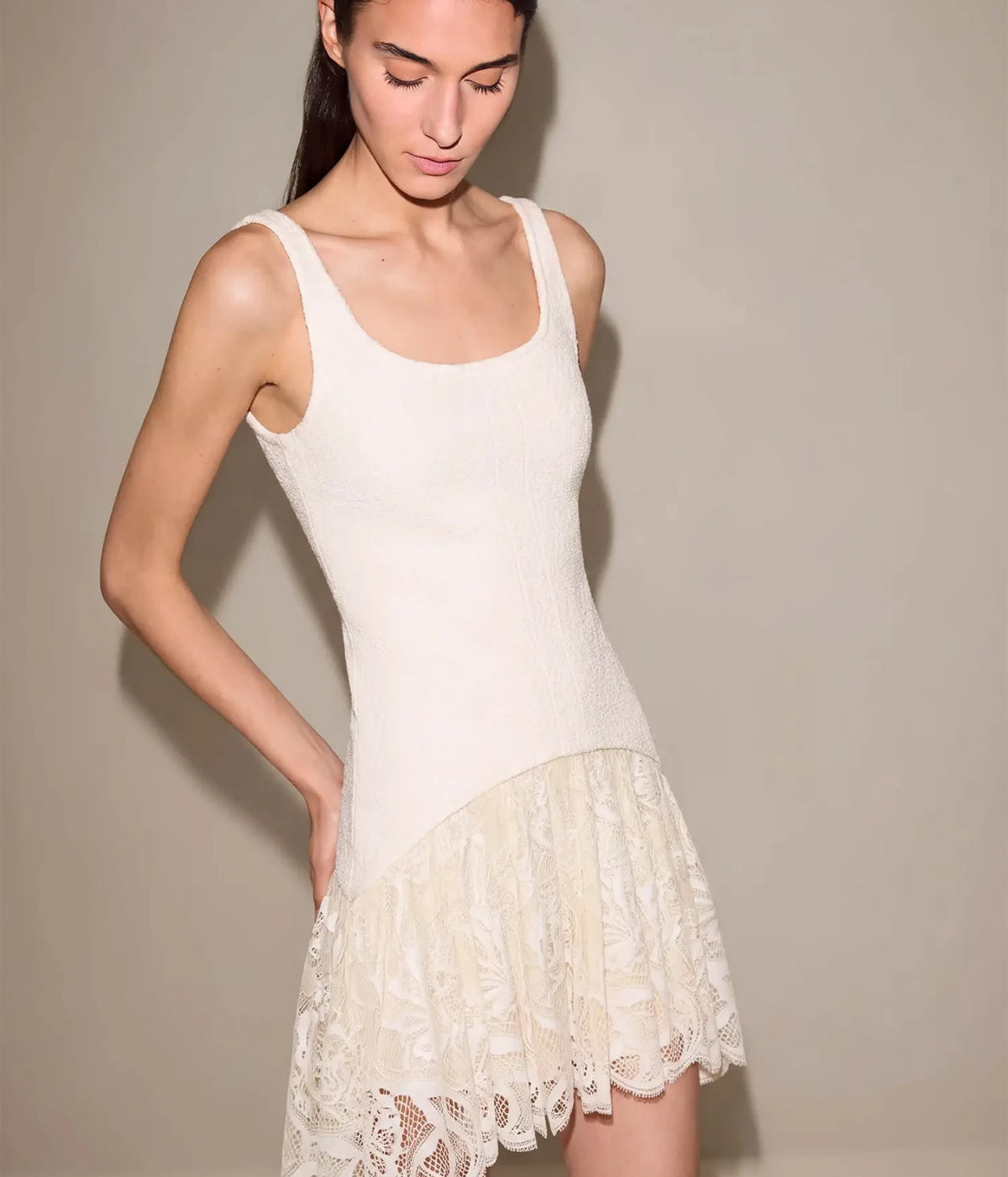 Three-quarter front view of a model wearing the Alexis Pomela Mini Dress In Ivory with a hand on her hip, showing the scoop neckline, textured fitted bodice and asymmetrical lace hem