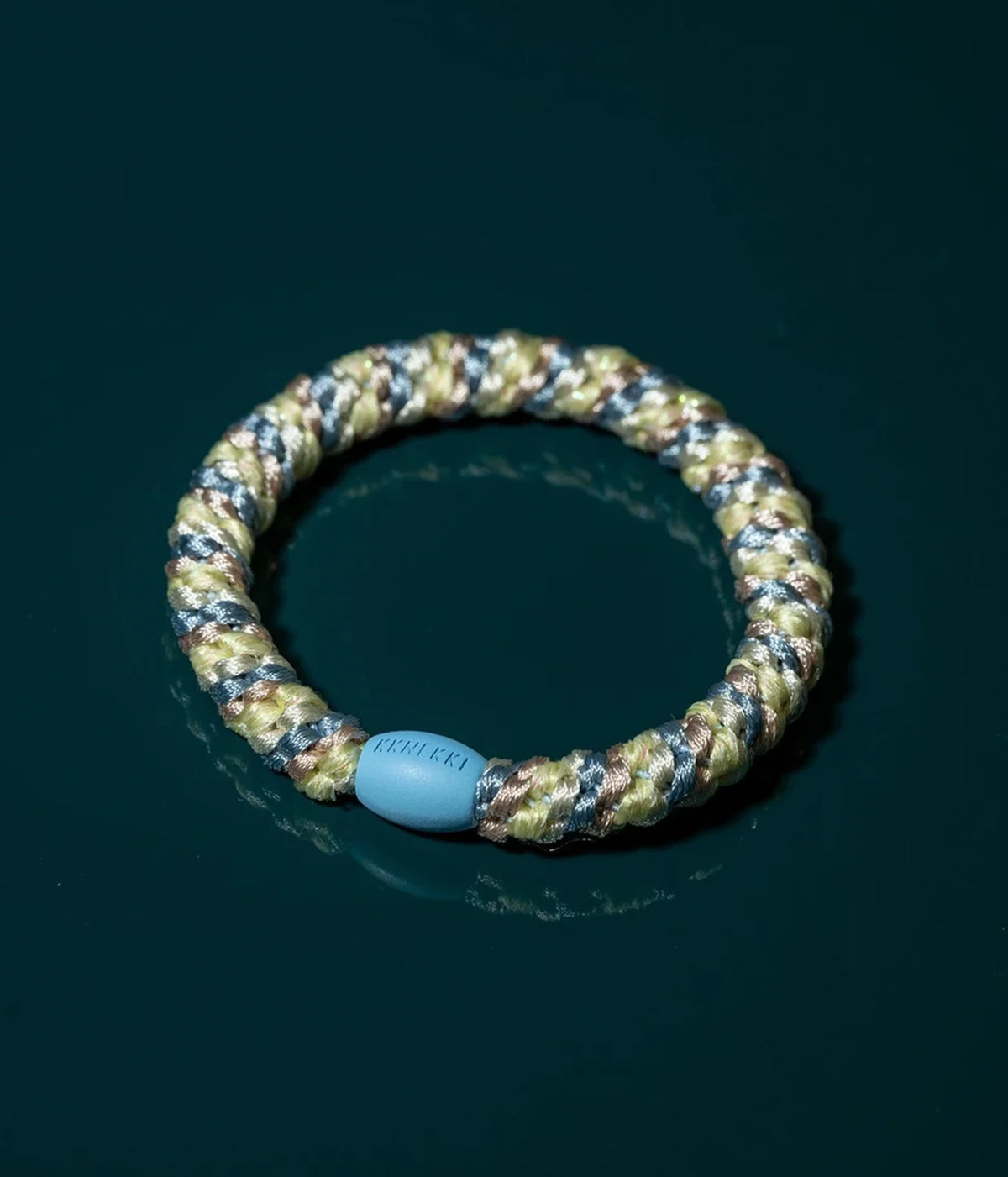 A braided elastic bracelet with a camouflage pattern of olive green, tan, and dark gray, featuring a light blue oval bead with the brand name 'KKNEKKI' on it