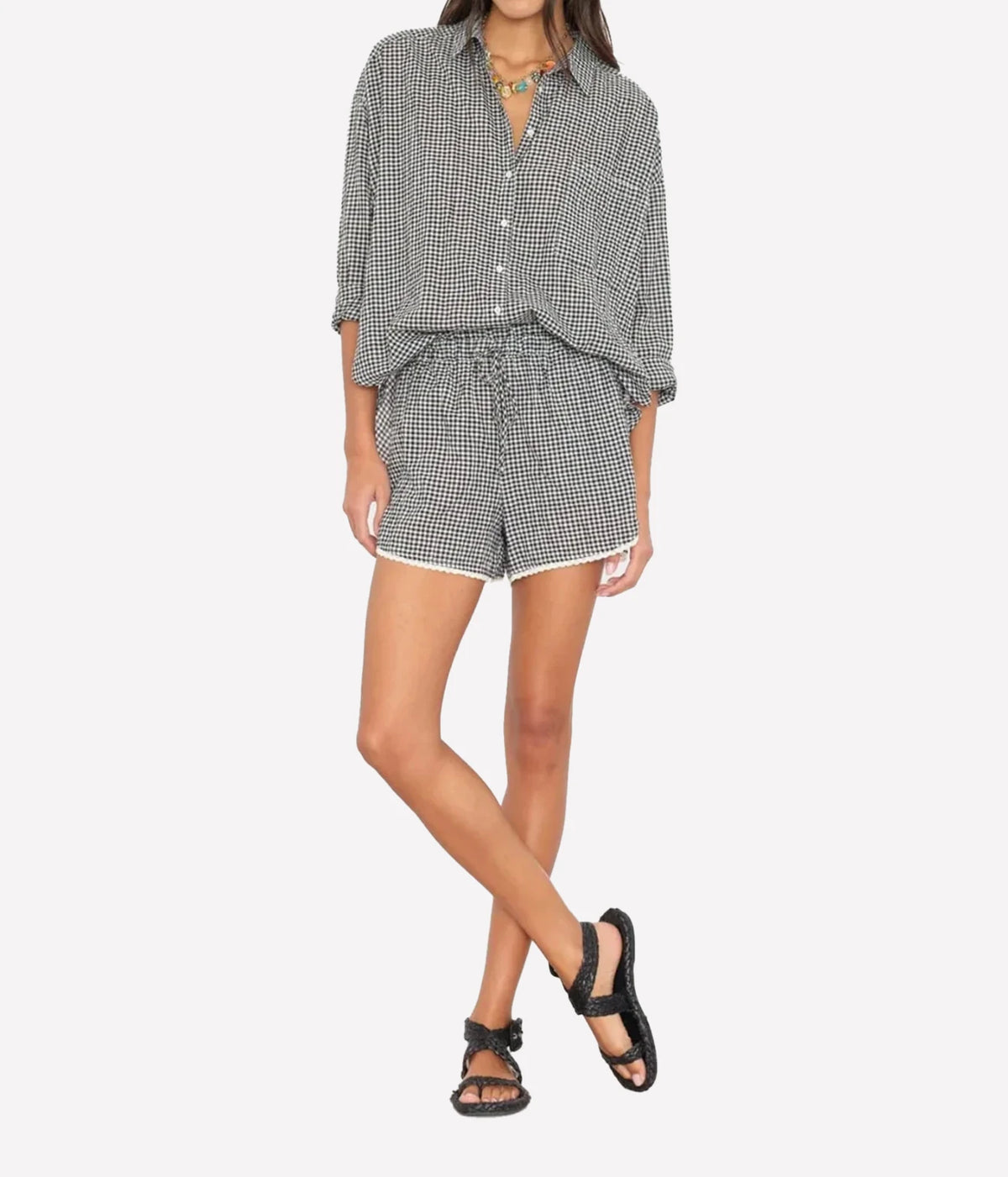 Model wearing the Xirena Starla Short in Black Sand with a matching gingham oversized button-up shirt with rolled sleeves, layered necklaces and black strappy sandals