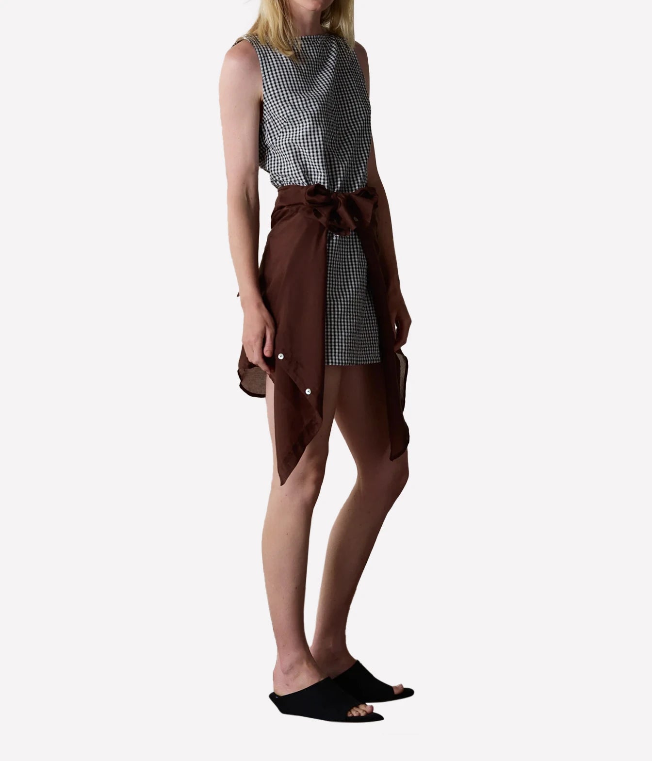 A model wearing a sleeveless black and white gingham top paired with a brown shirt-style wrap skirt tied at the waist, styled with black slide sandals against a plain light gray background.