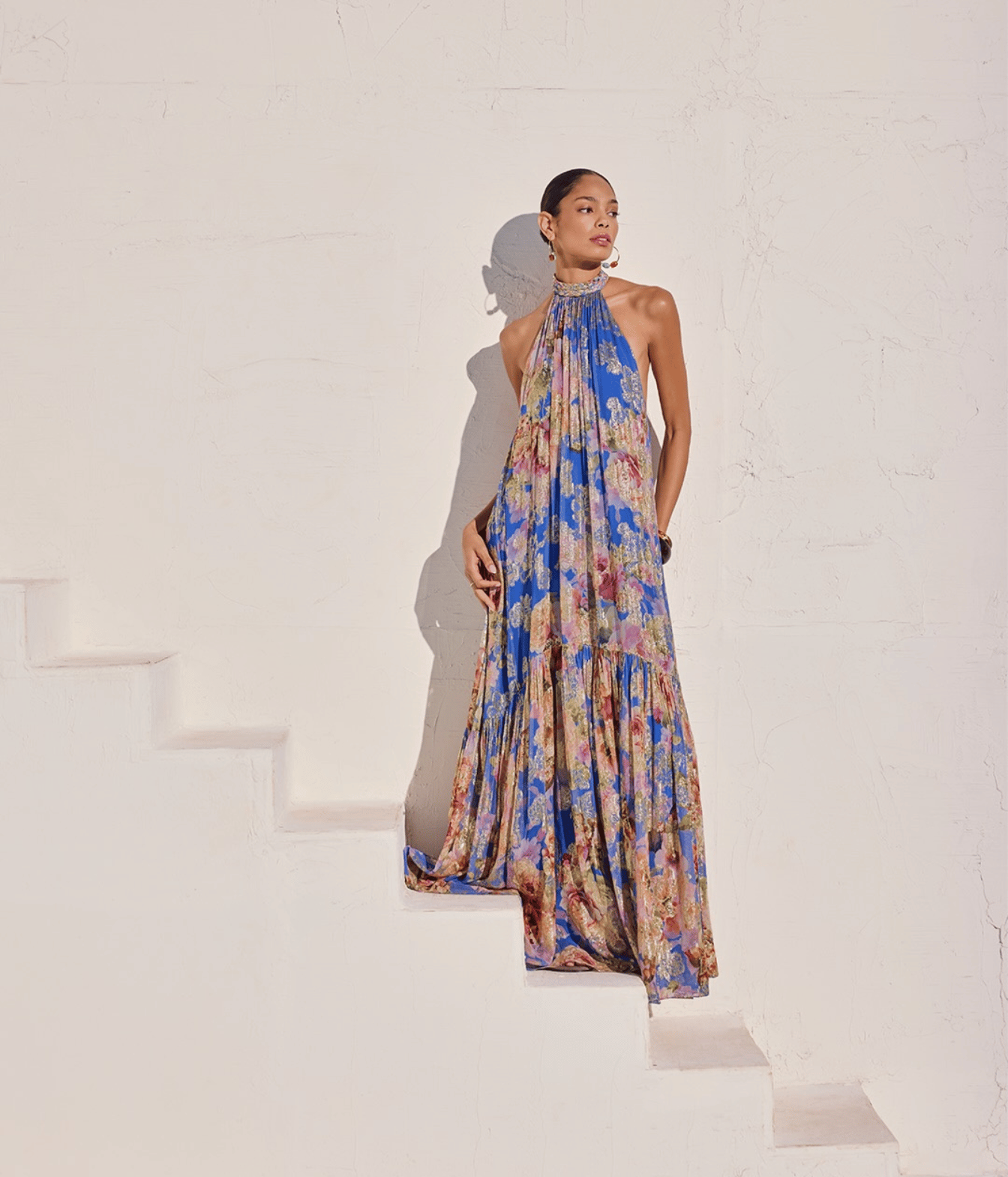 A model wearing a halter-neck, tiered maxi dress with a vibrant blue, pink, and beige floral pattern, posing on white stairs against a textured white wall.