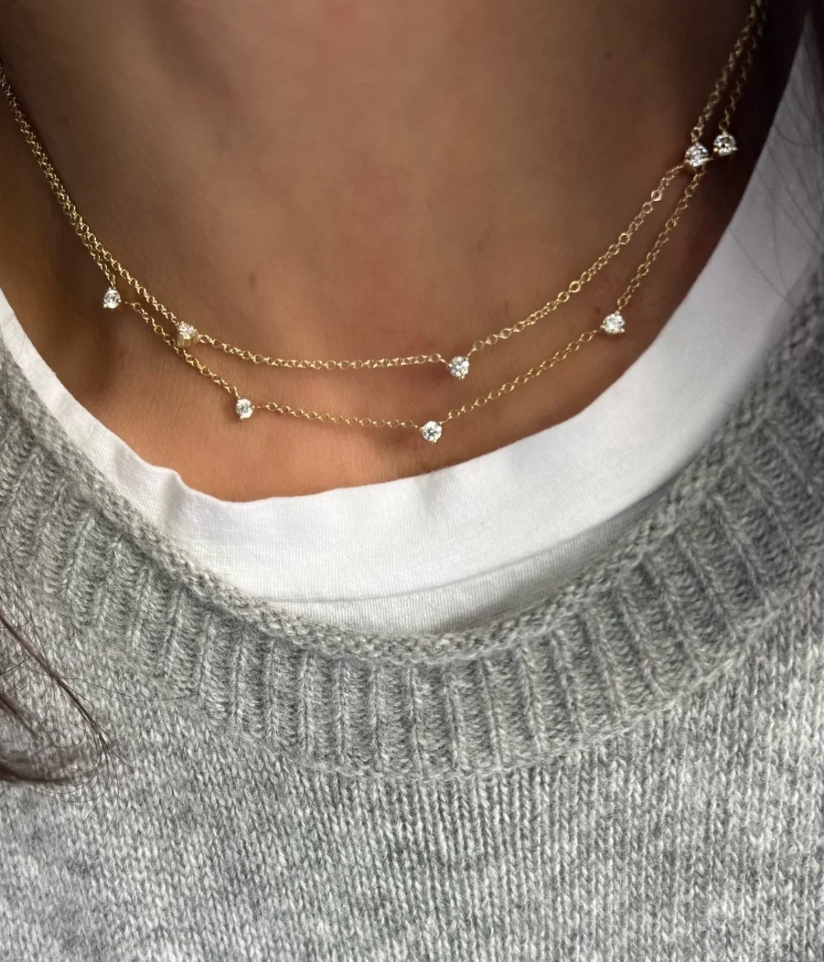 Close-up of a model wearing the Shylee Rose 5 Floating Diamond Necklace In 14K Yellow Gold layered with a second fine gold diamond chain, worn over a white T-shirt and grey knit jumper
