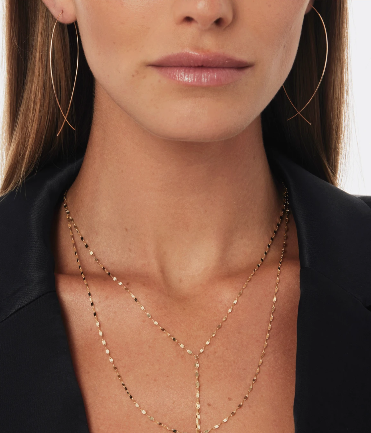 Close-up of a model wearing the LANA Small Upside Down Hoops in Yellow Gold sweeping past the jawline, styled with a black open-collar blazer and layered gold chain necklaces including a lariat