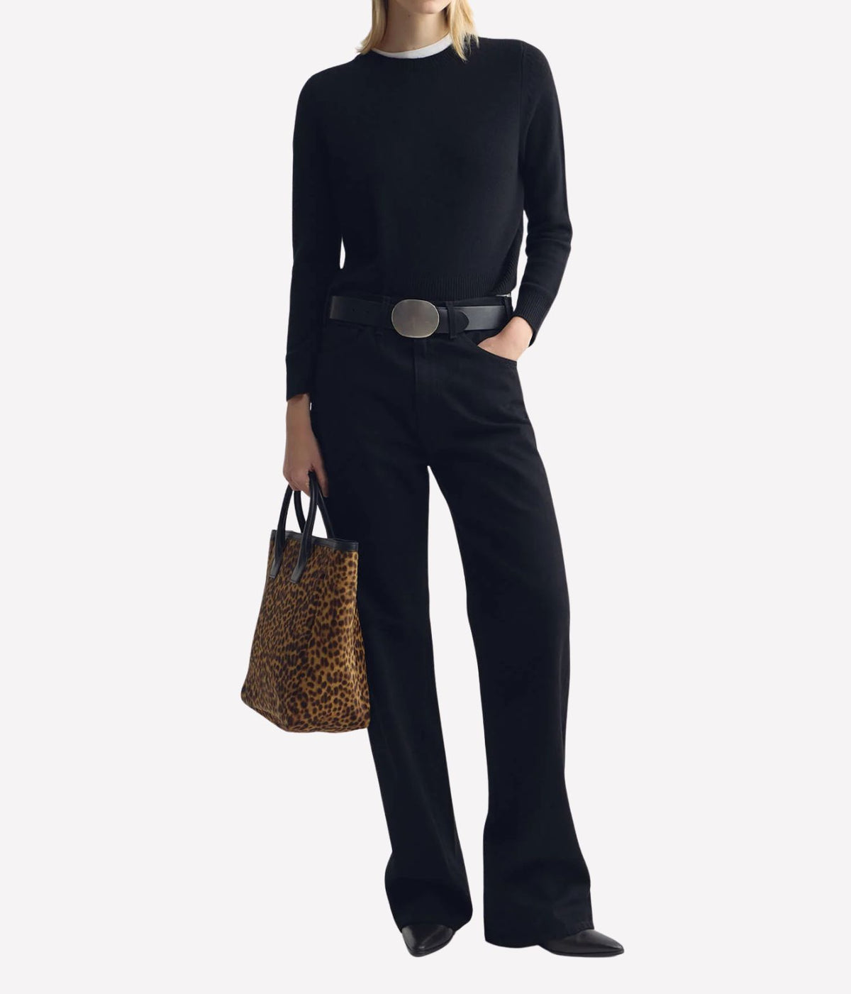 A model wearing a black long-sleeve top, black wide-leg pants, and a black belt with a silver buckle, holding a leopard-print tote bag against a light gray background.