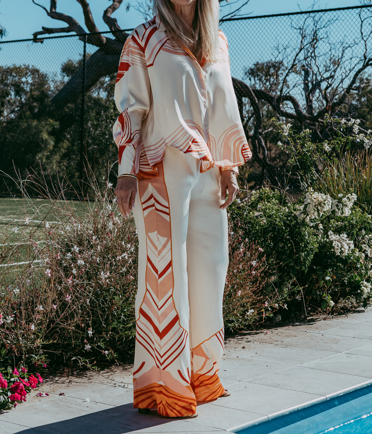 A woman wearing a matching two-piece lounge set with a cream base and orange geometric patterns, standing outdoors next to a swimming pool.