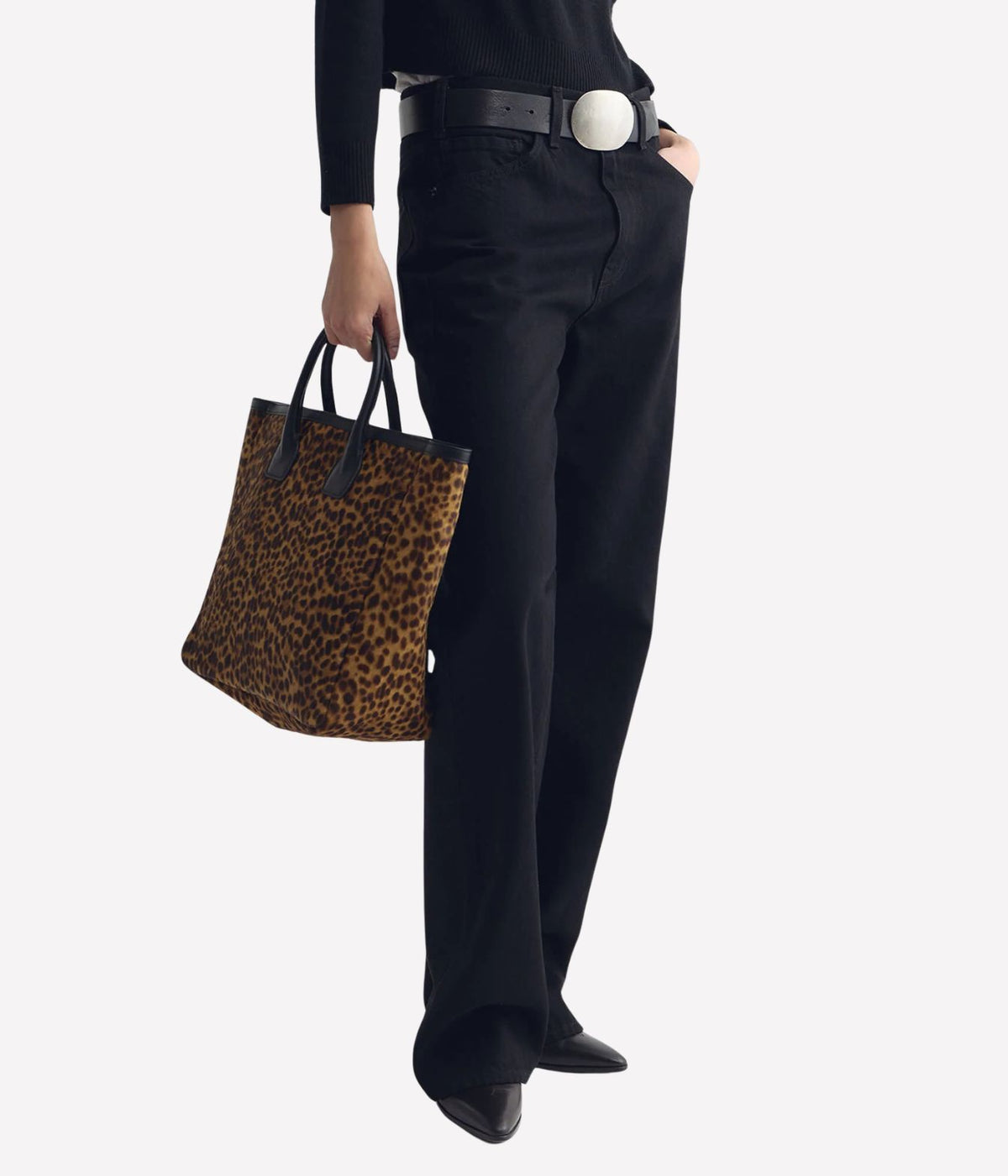 A person wearing black pants, a black top, and a black belt with a large silver buckle, holding a leopard-print tote bag against a light gray background.