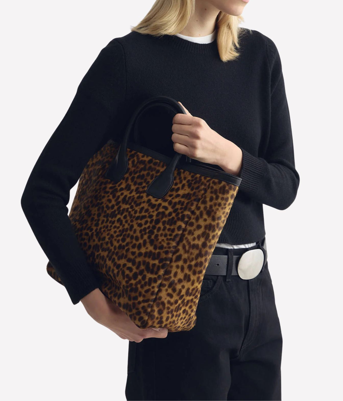A model wearing a black knit sweater and black pants holds a leopard-print tote bag with black handles against a light gray background.